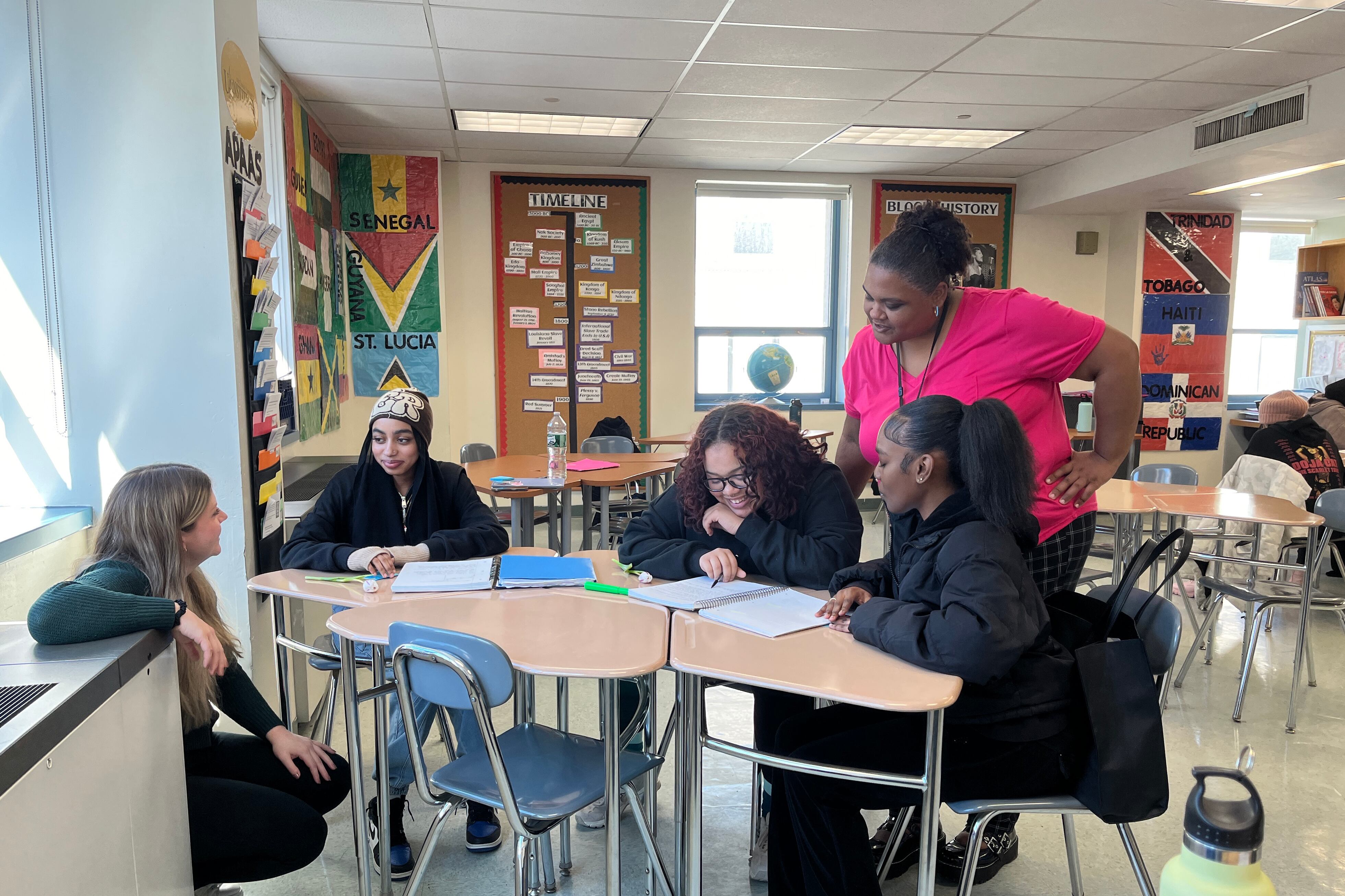 Two adults and three students stand and sit around a cluster of wooden desks in a classroom with decorated walls in the background.