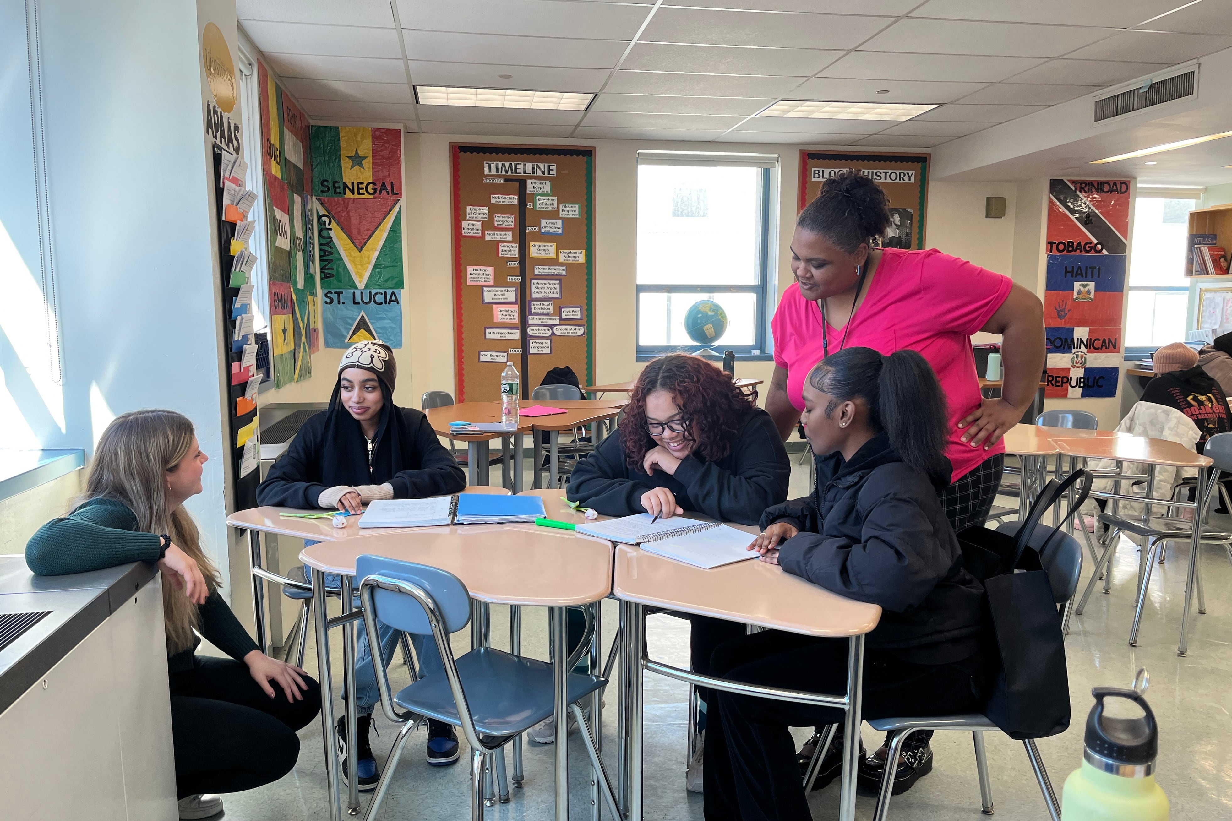 Two adults and three students stand and sit around a cluster of wooden desks in a classroom with decorated walls in the background.