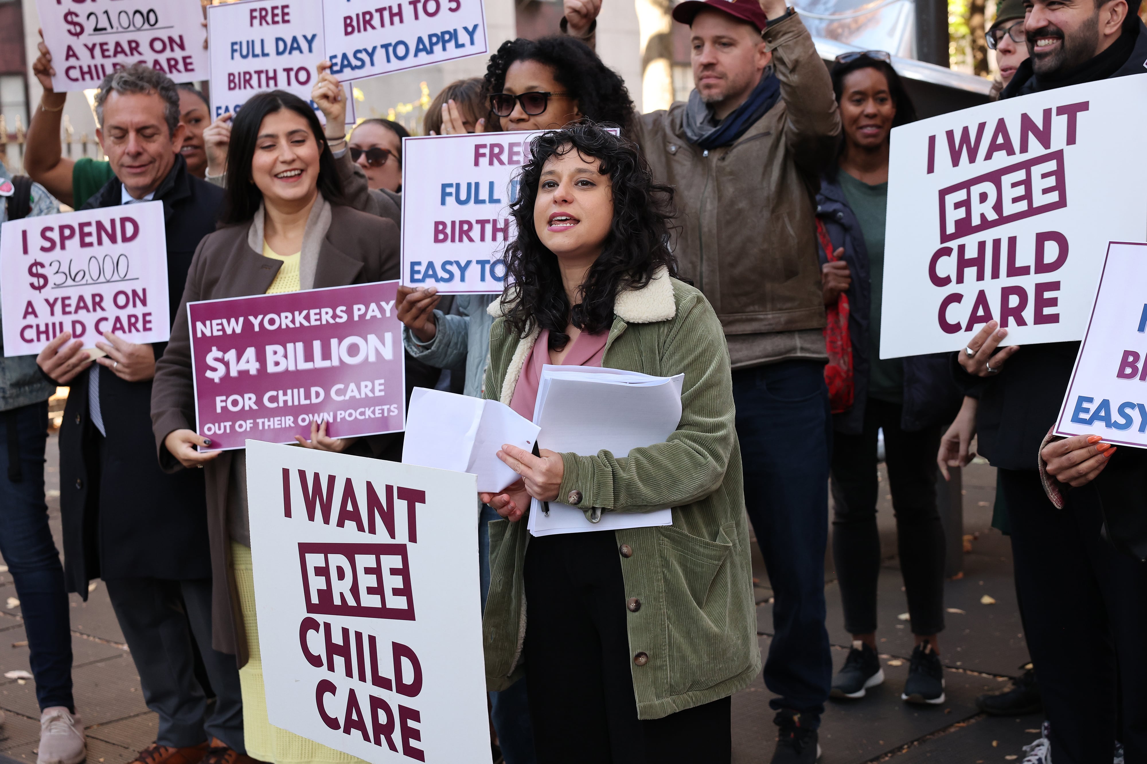 A group of people hold signs in a group outside while one woman stands in the front speaking.