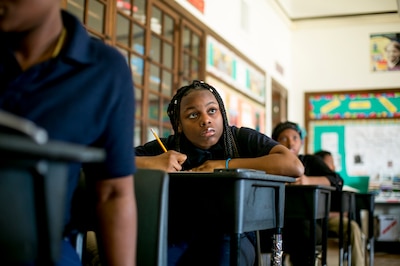 One young girl is looking up from her school desk and holding a pencil while there are several students sitting in front and behind her in a row of desks.