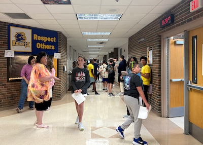 A photograph of a group of middle school students walking in a hallway of a school with a couple of adults scattered throughout.