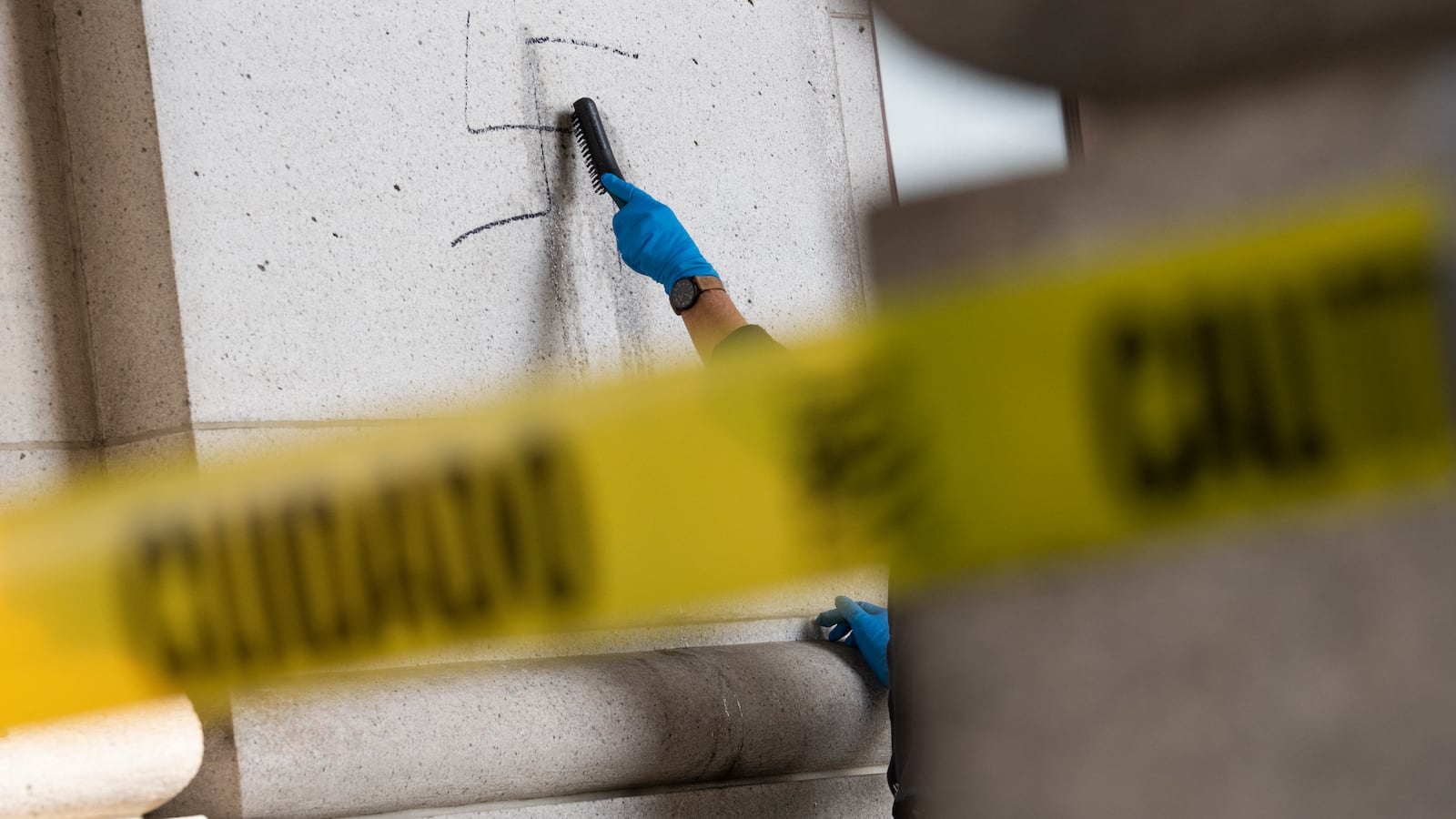 A person works to remove a swastika from an exterior wall. There is caution tape in the foreground.