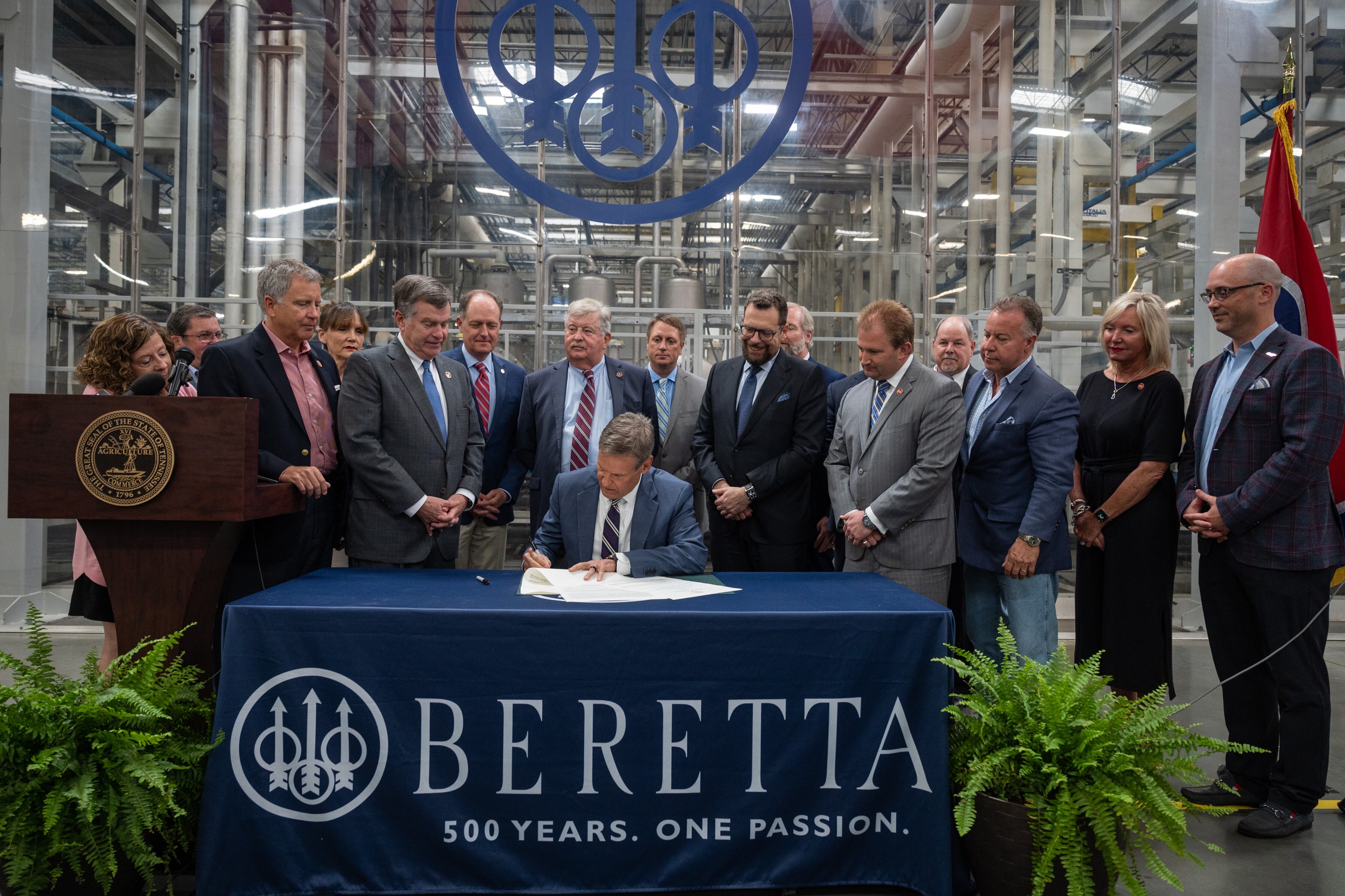 A man wearing a suit signs a document inside of a large manufacturing plant and surrounded by a dozen other people wearing suits and dresses.