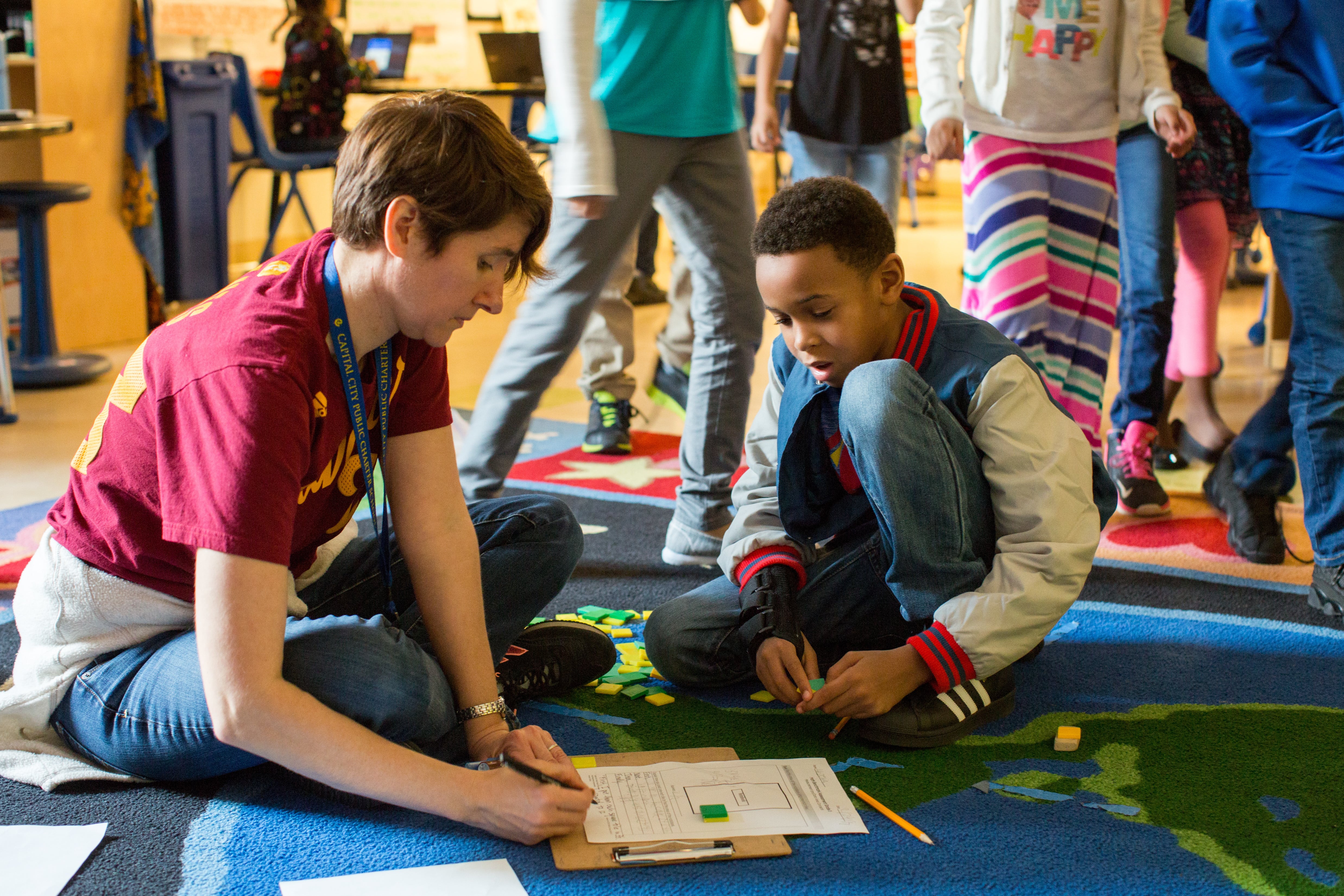 A female teacher wearing a red shirt sits on a carpet and helps a male student in third grade with a math assignment.