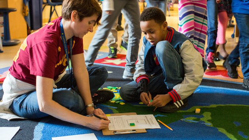 A female teacher wearing a red shirt sits on a carpet and helps a male student in third grade with a math assignment.