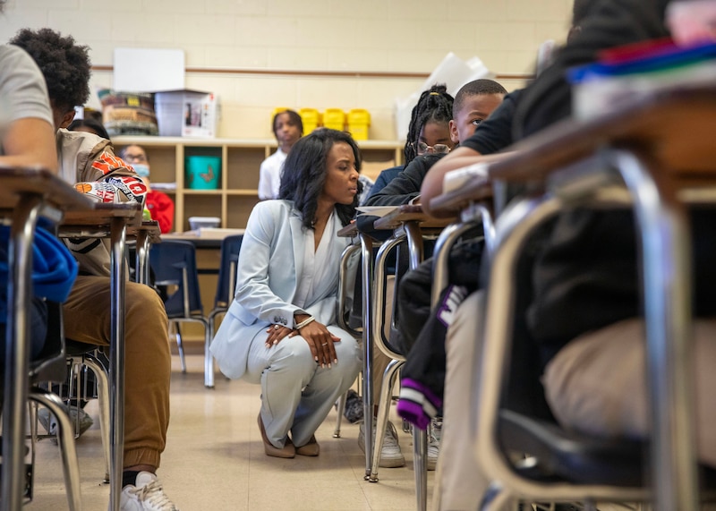A woman with short dark hair and wearing a light blue suit kneels next to a desk with a student between two rows of desks in a classroom.