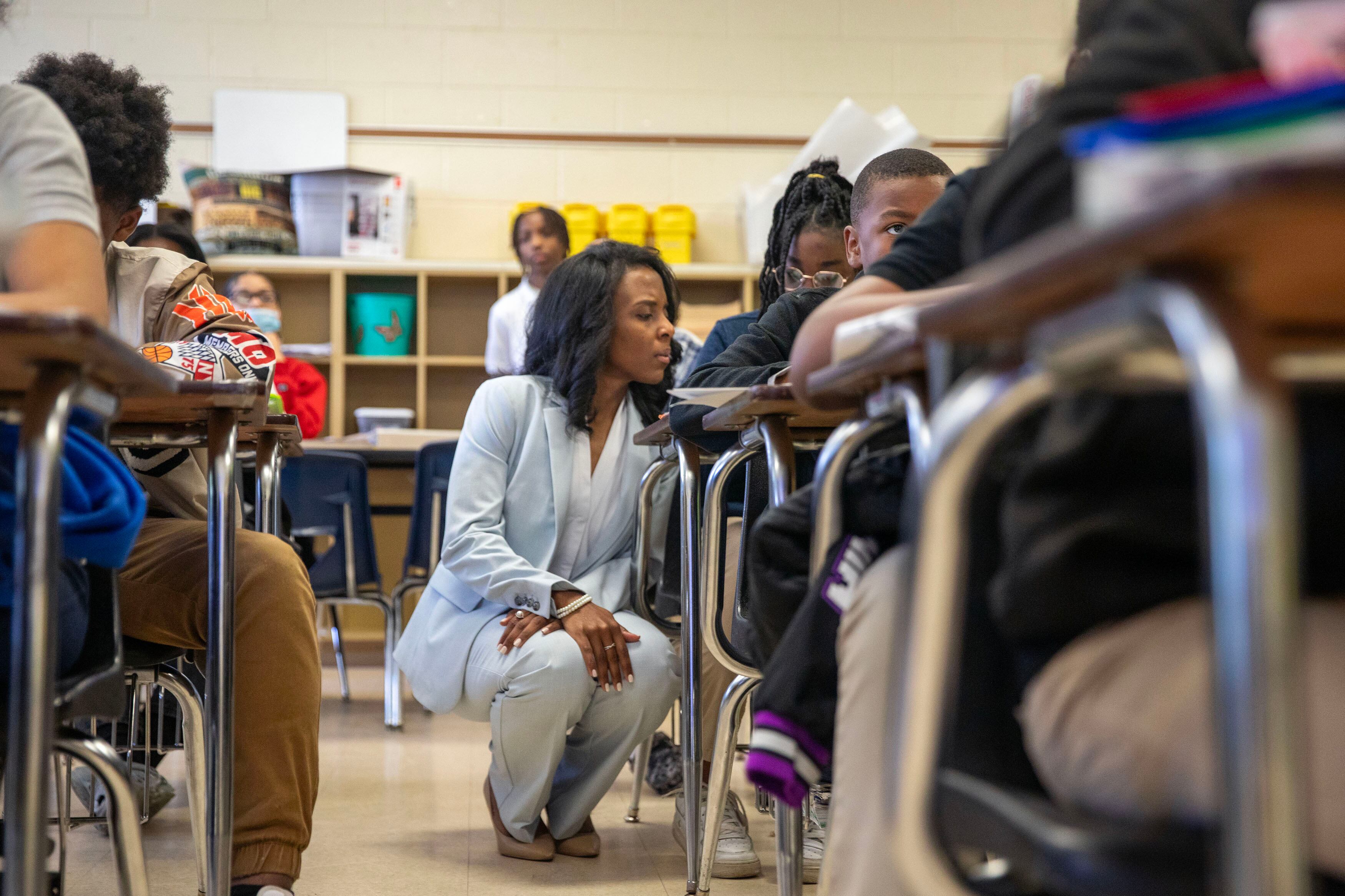 A woman with short dark hair and wearing a light blue suit kneels next to a desk with a student between two rows of desks in a classroom.