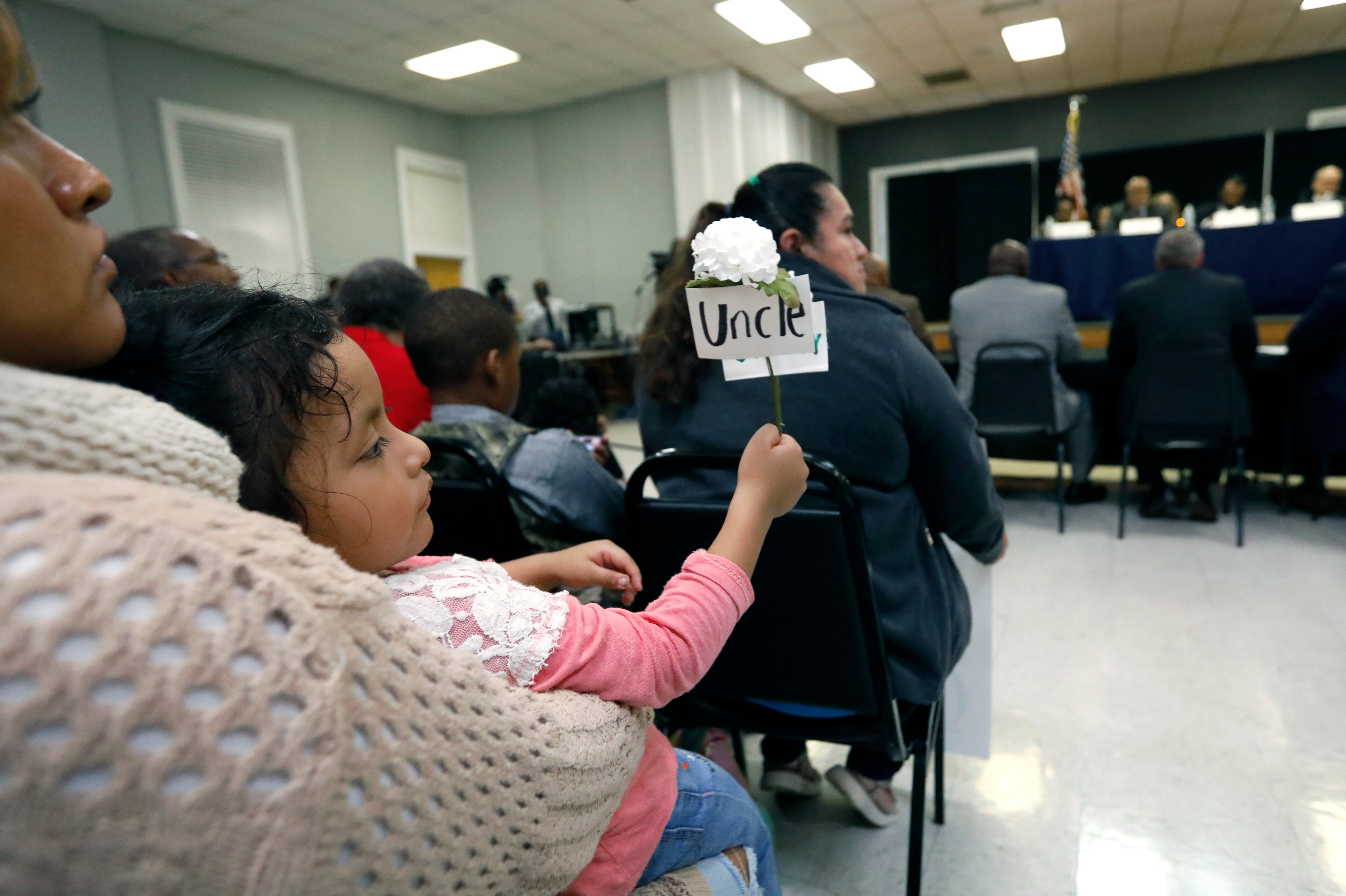 A little girl sits in her mom's lap holding a white carnation with a tag attached that says "UNCLE." They're seated in a hearing room with many other people.