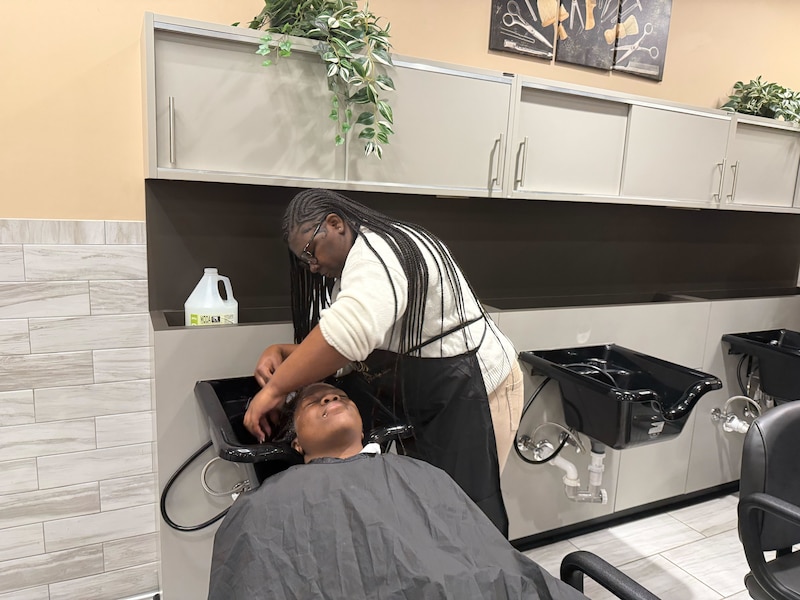 A woman with a black apron uses a hose to wash the hair of a woman lying back on a chair.