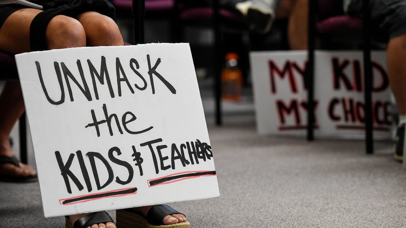 A person holds a sign that reads “unmask the kids & teachers” during a protest at a board of education meeting.
