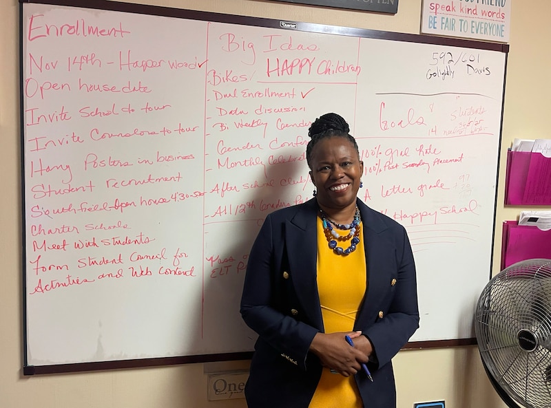 A photograph of a Black woman in a yellow dress and blue blazer standing for a portrait in front of a dry erase board with red writing across the whole thing in a classroom.