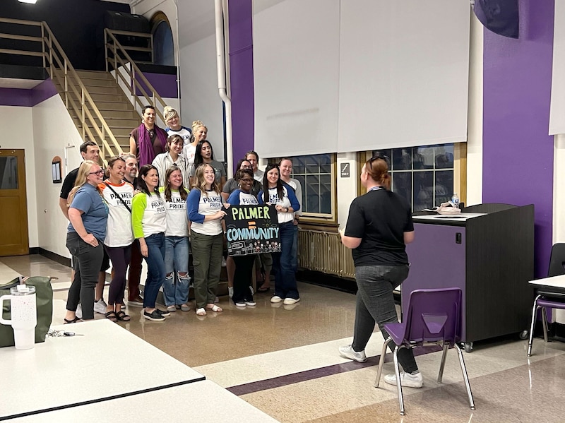 A group of people holding a sign and posing for a photo in front of a person making the photo in a school common area.