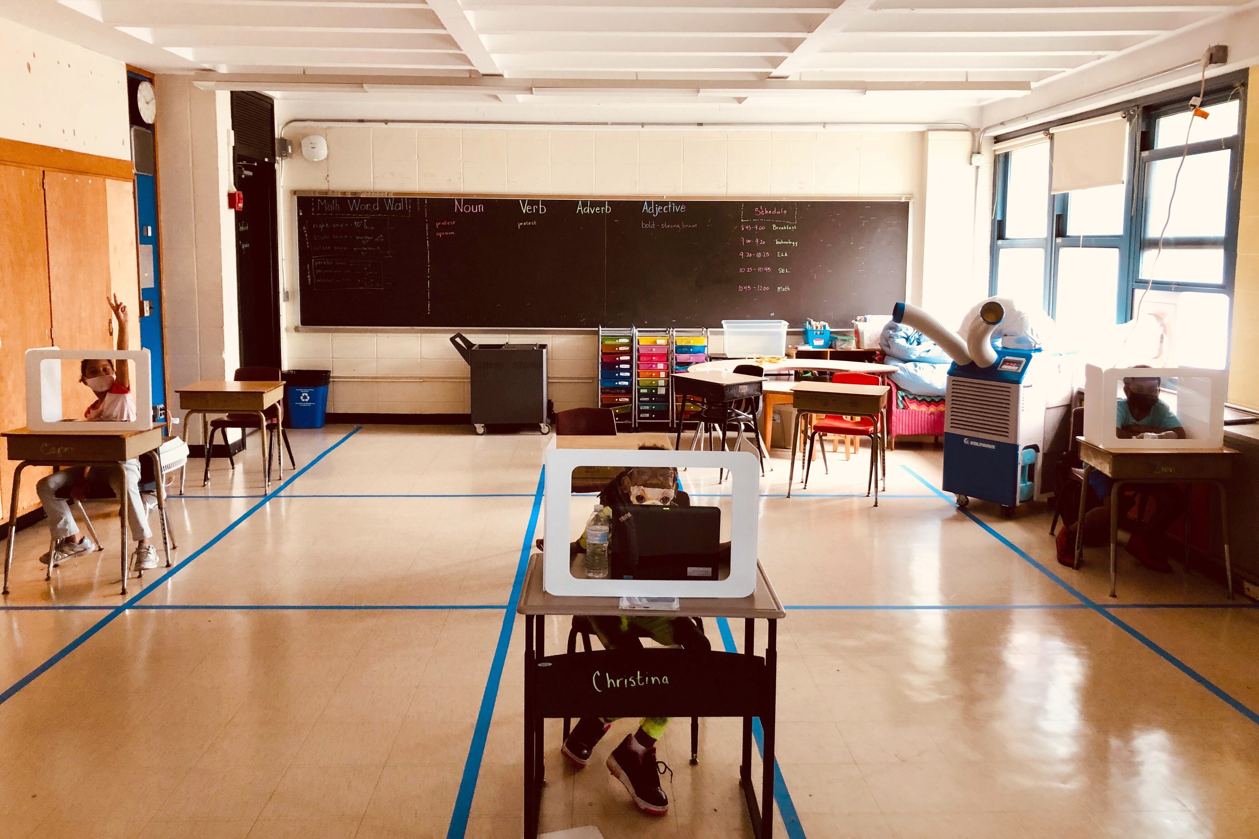 Three masked elementary students sit behind clear shields placed on widely separated desks in a classroom with a portable air-conditioning unit. The blackboard has a “Math Word Wall” and a space with “Noun Verb Adverb Adjective” written, as well as the class schedule.