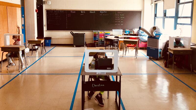 Three masked elementary students sit behind clear shields placed on widely separated desks in a classroom with a portable air-conditioning unit. The blackboard has a “Math Word Wall” and a space with “Noun Verb Adverb Adjective”  written, as well as the class schedule.
