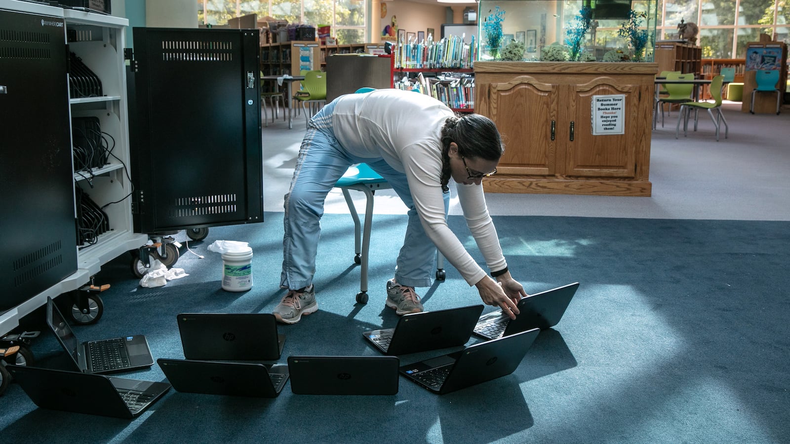 A teacher wearing jeans and a white shirt cleans a bunch of Chromebook laptop computers/