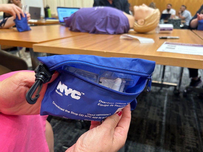A person holds a small blue bag that has white letters on it and there is a practice doll on a table in teh background.