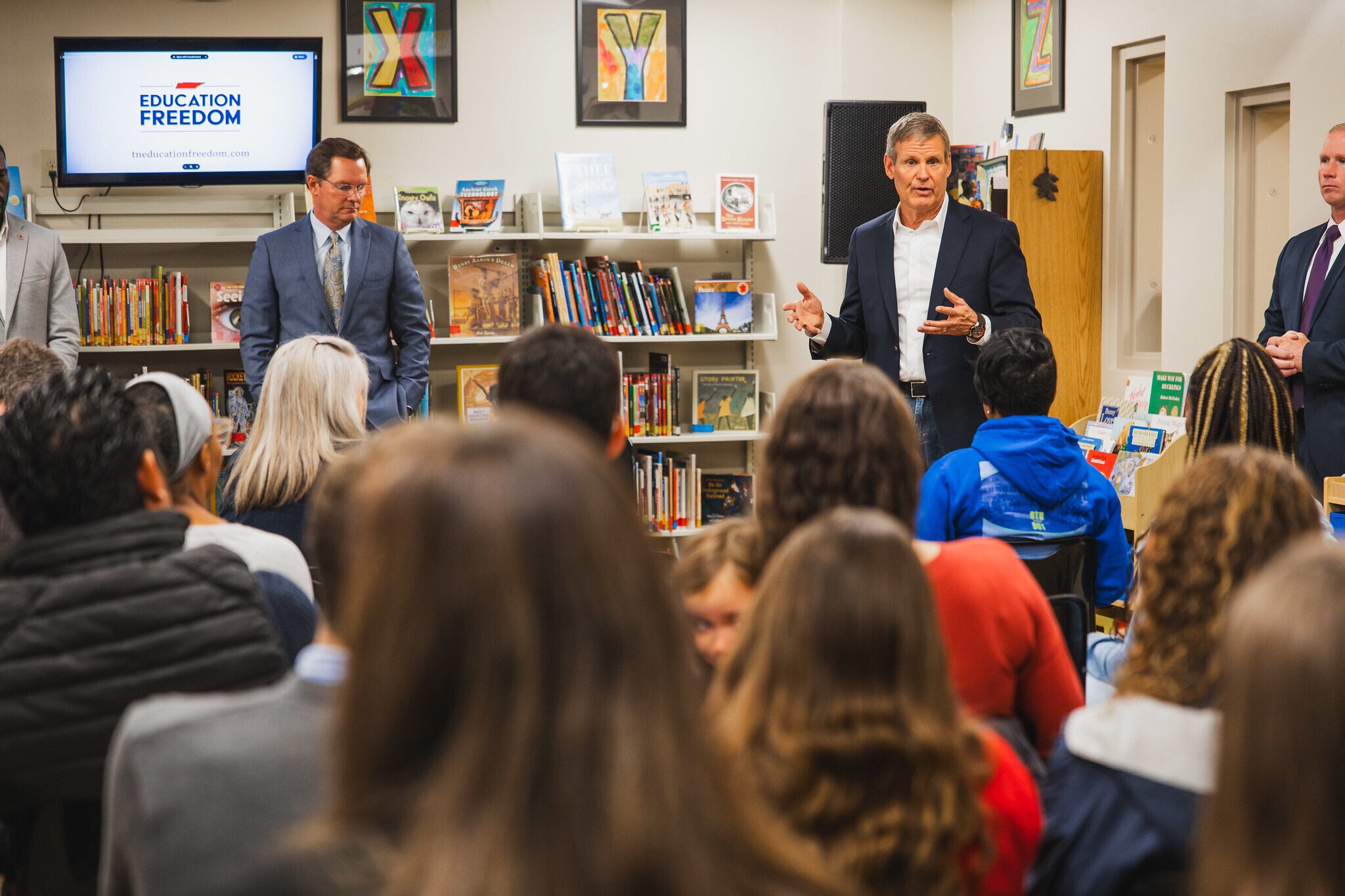 Two adults speak in front of a crowd of people sitting in chairs in rows in a classroom.