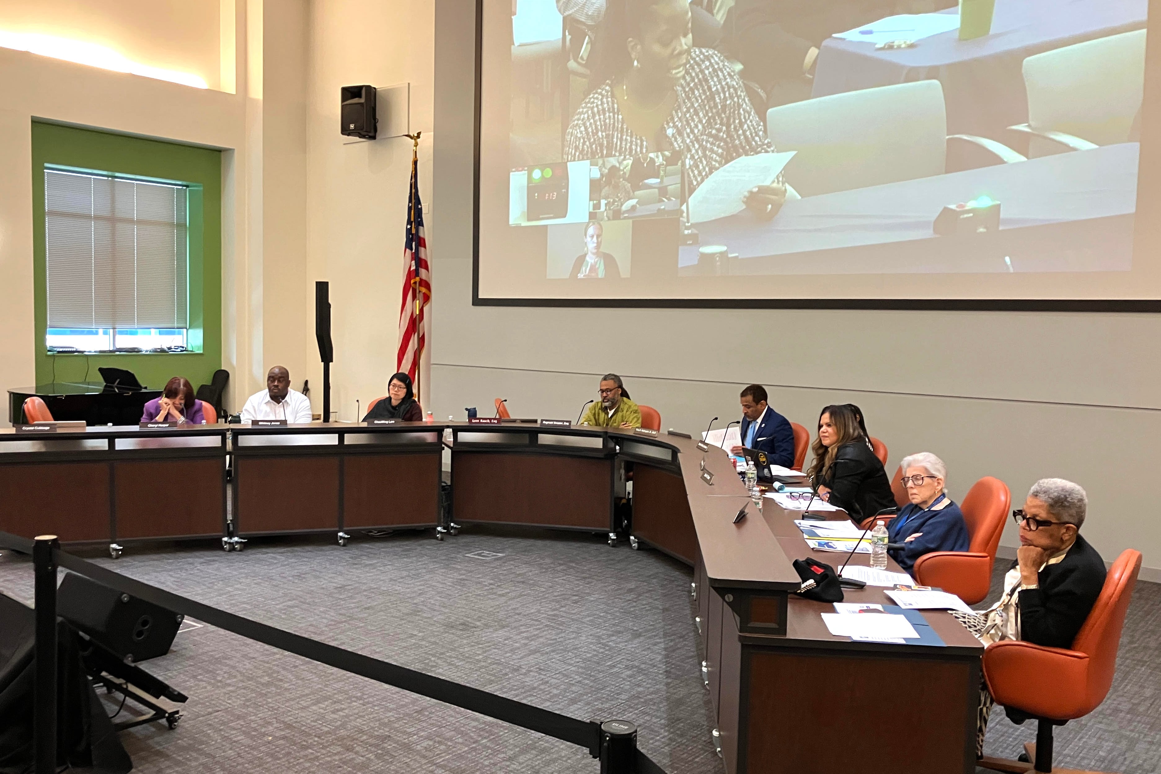 Men and women sit at a long desk in front of a screen.