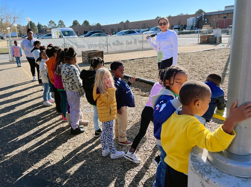 Young students are in a line outside with two adults on each end of the line. Cars in a parking lot and a building are in the background.