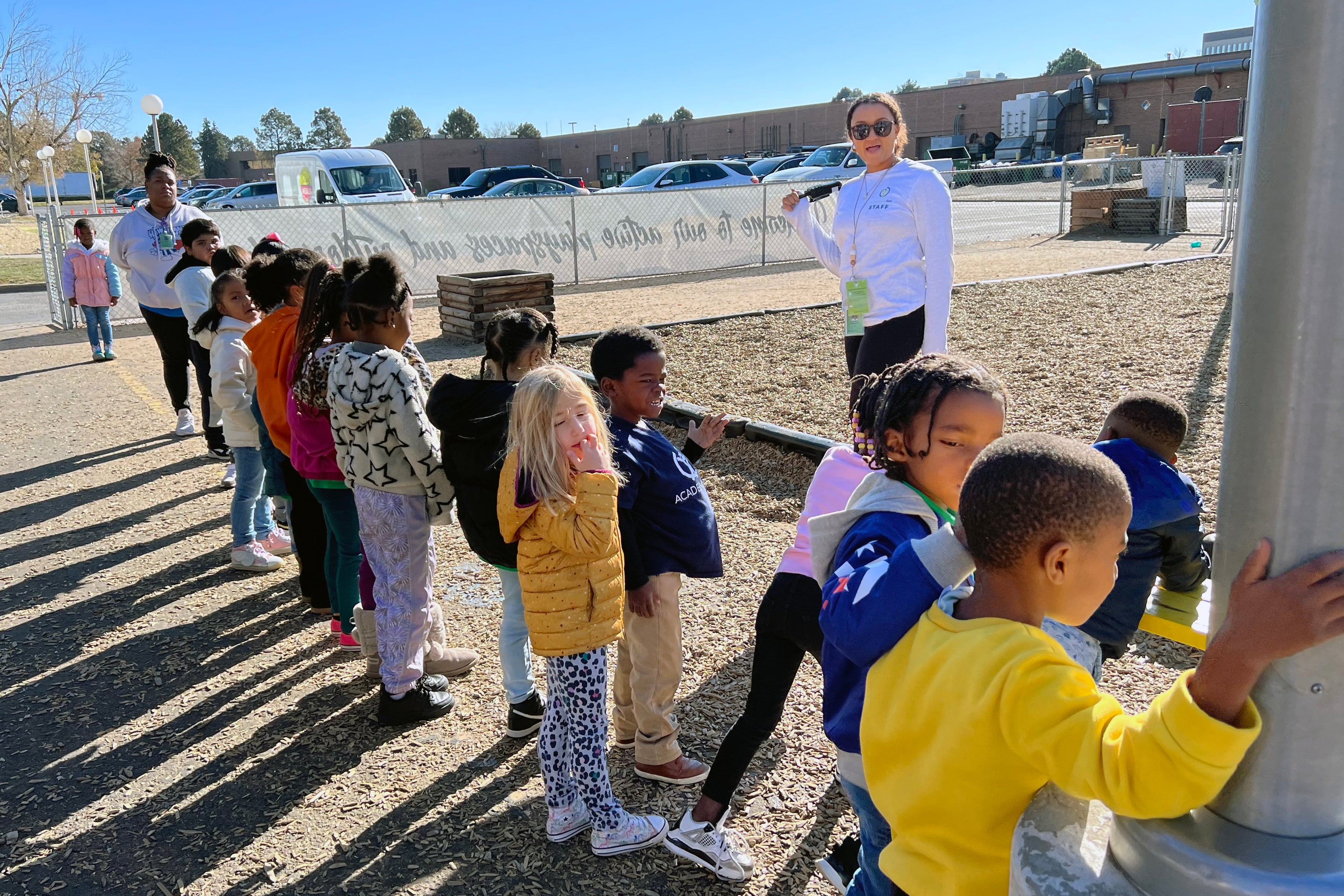 Young students are in a line outside with two adults on each end of the line. Cars in a parking lot and a building are in the background.