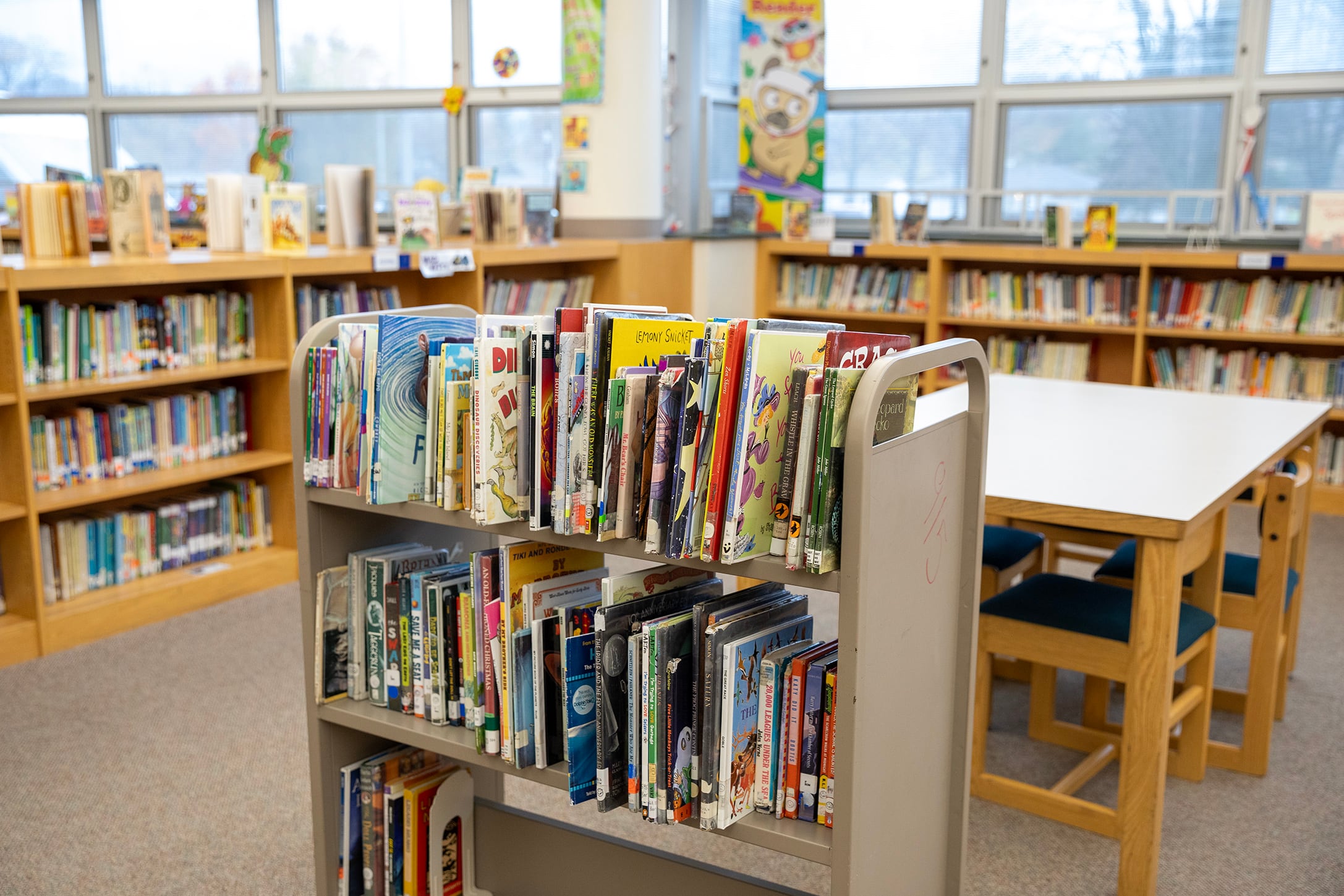 A book cart full of books sits in the middle of a library with half shelves lining the room in the background.