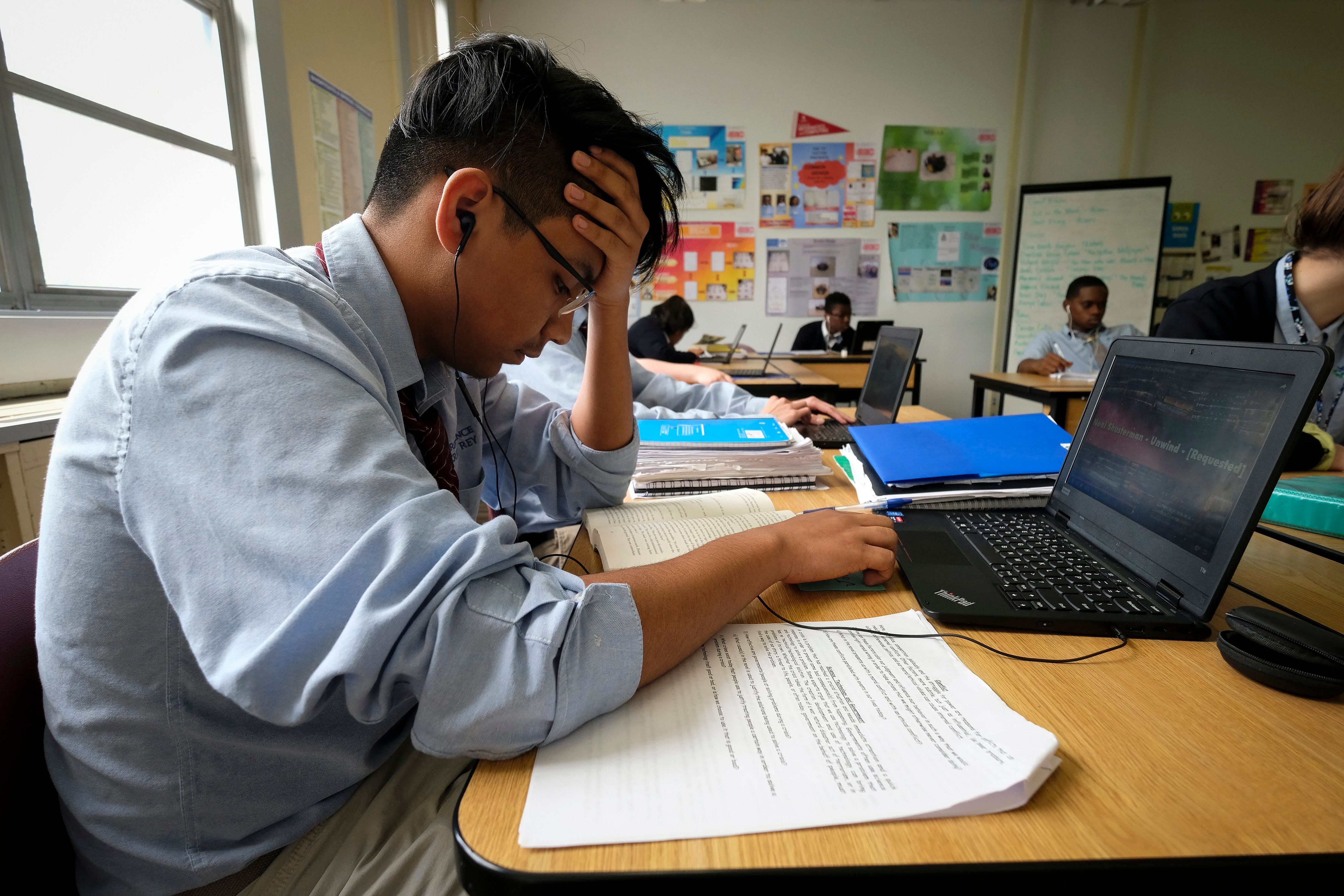 A photograph of a high school student with his head in his hand reading from a notebook and working from a computer.