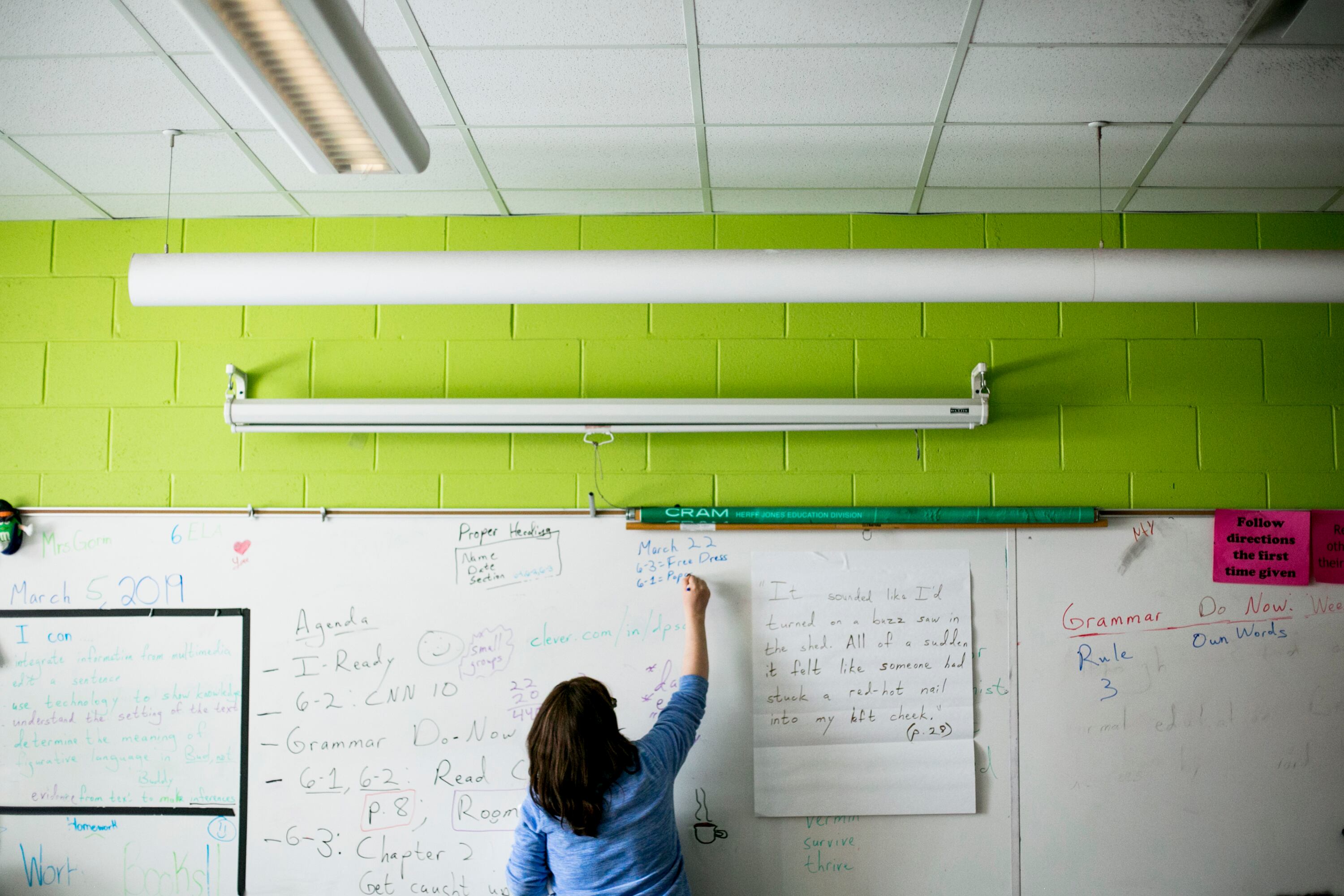 An adult teacher with short dark hair and wearing a blue shirt writes on a dry erase board in a classroom.