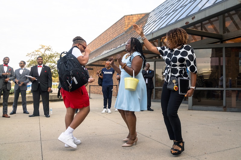 A student wearing a red backpack greets two adults outside of a school building with a group of adults lining the background.