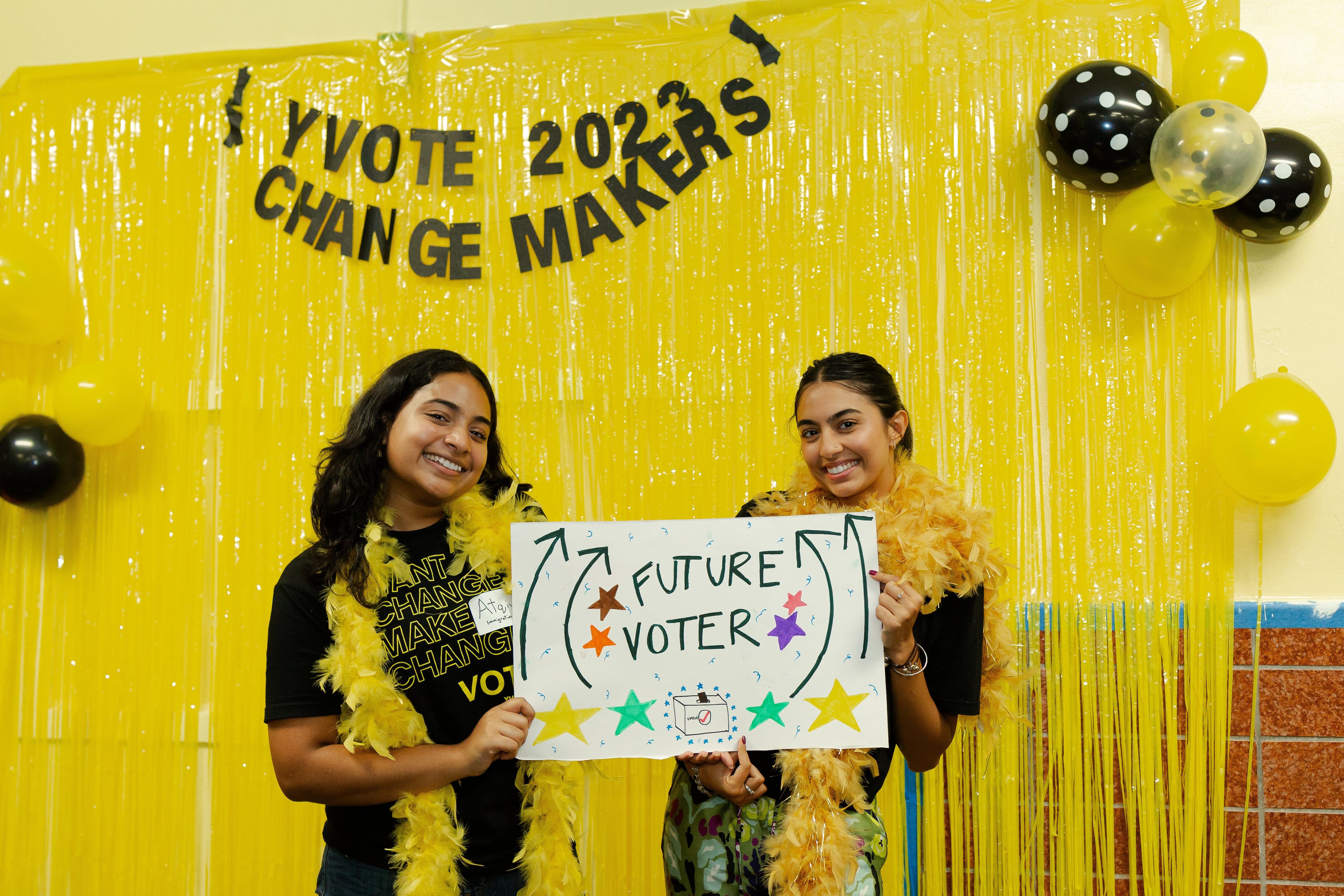 Two young women pose in front of a yellow curtain holding a sign saying, "Future Voter."