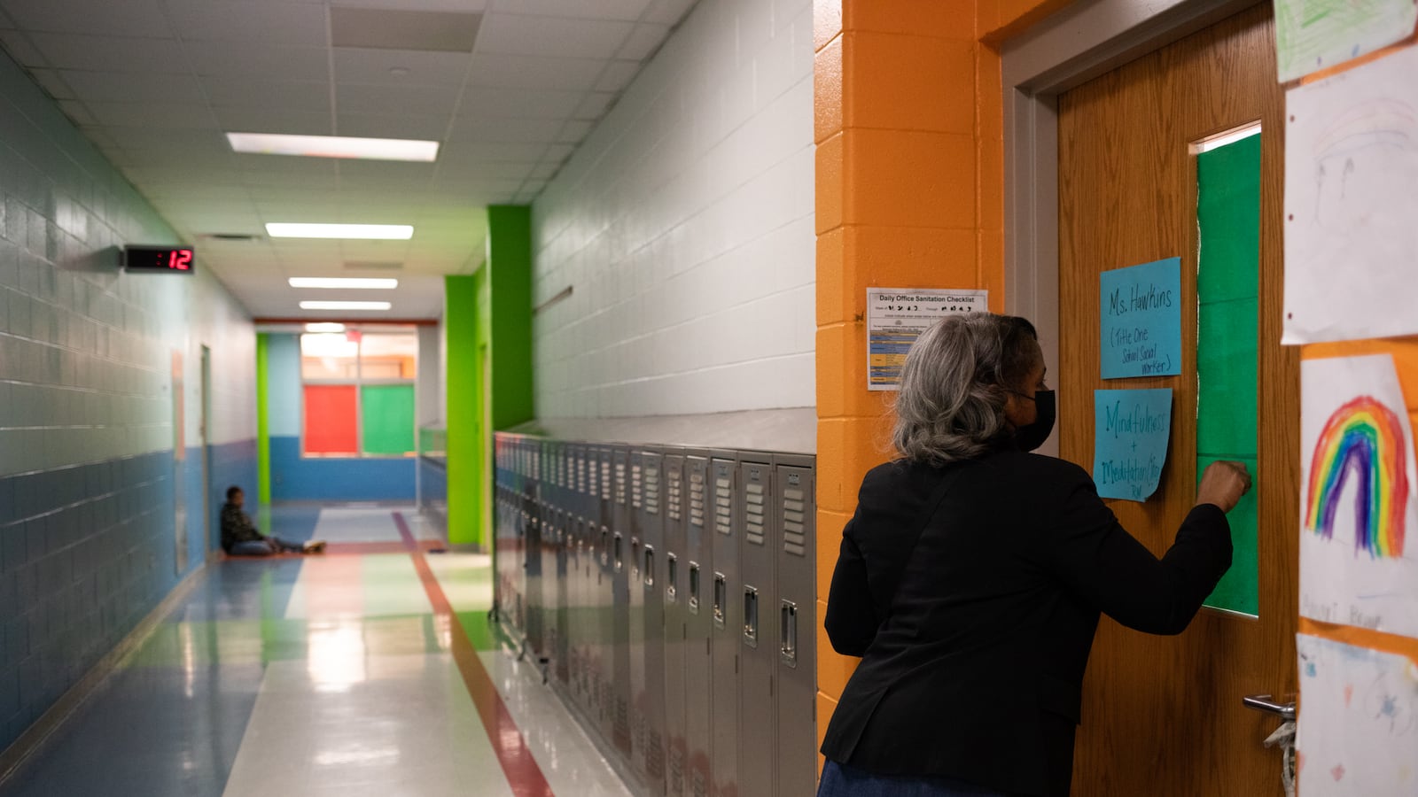 A woman knocks on a classroom door in a mostly empty classroom. There is a boy in the background sitting on the floor.