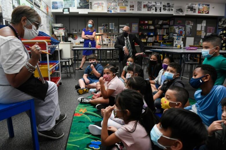 A teacher reads a book to students sitting on a rug. The teacher and students are wearing masks.