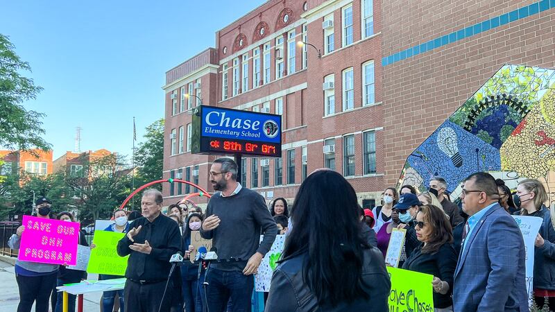 A man stands at a microphone and speaks in front of a crowd of parents and children outside of a school.