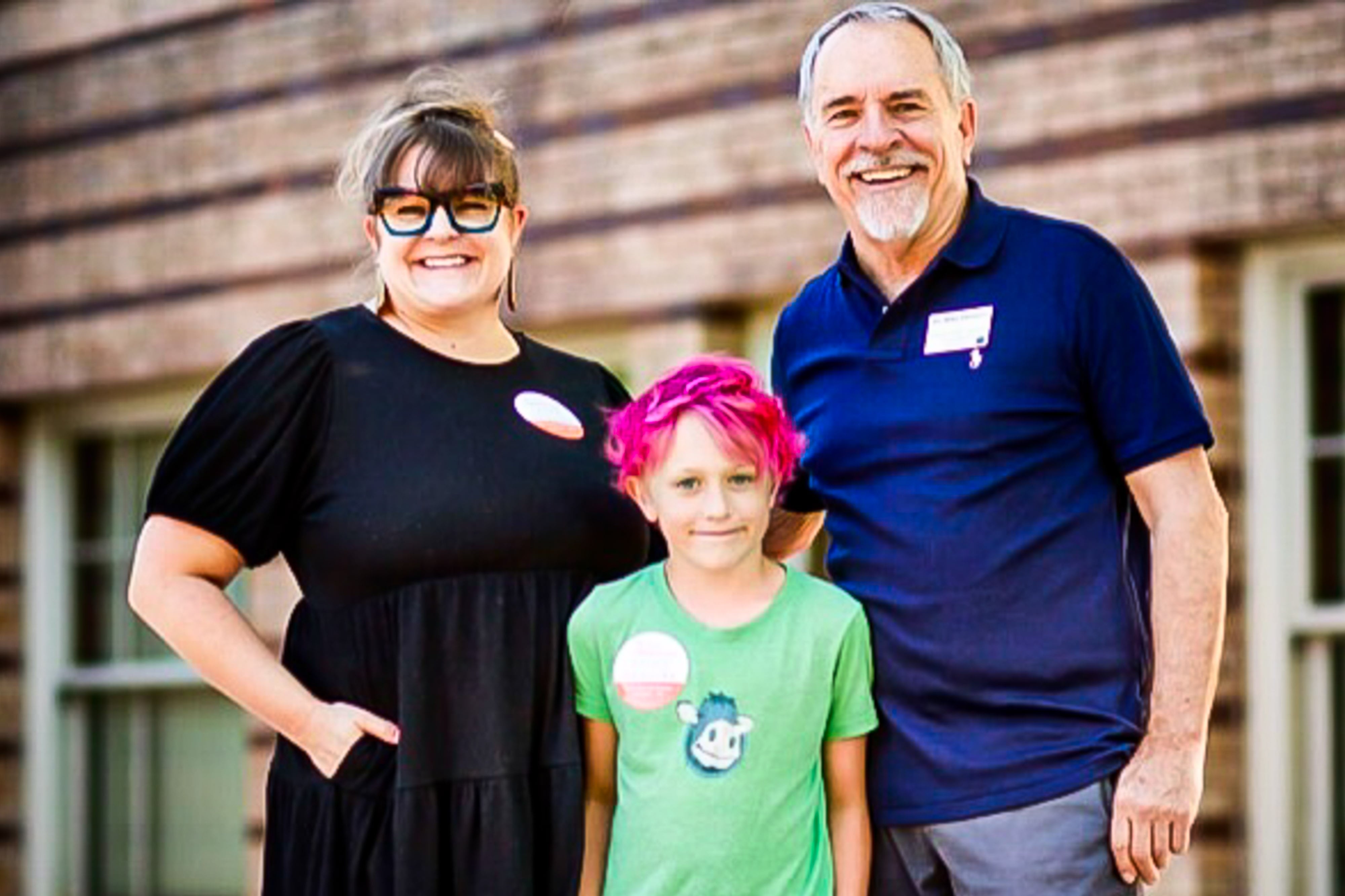 Denver school board candidate Mike DeGuire, in a blue polo shirt, stands in front of a wooden wall and windows, with his daughter, wearing a black dress, and his grandson in a lime green shirt and whose hair is dyed hot pink.