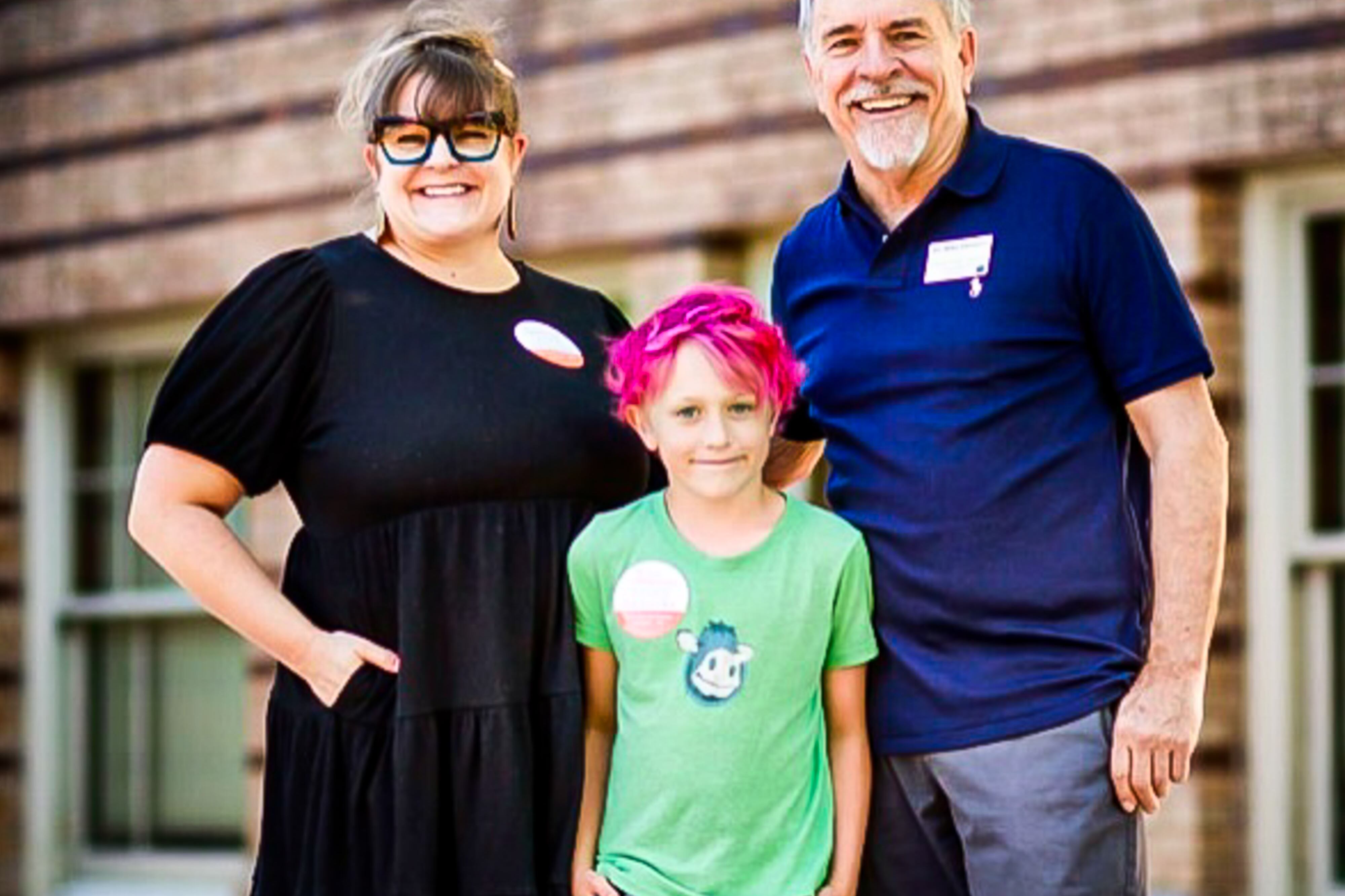 Denver school board candidate Mike DeGuire, in a blue polo shirt, stands in front of a wooden wall and windows, with his daughter, wearing a black dress, and his grandson in a lime green shirt and whose hair is dyed hot pink.