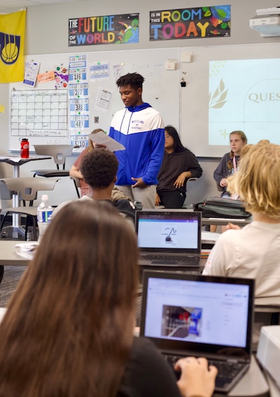 An eighth grade student stands up in the middle of a group of students sitting in chairs in a classroom.