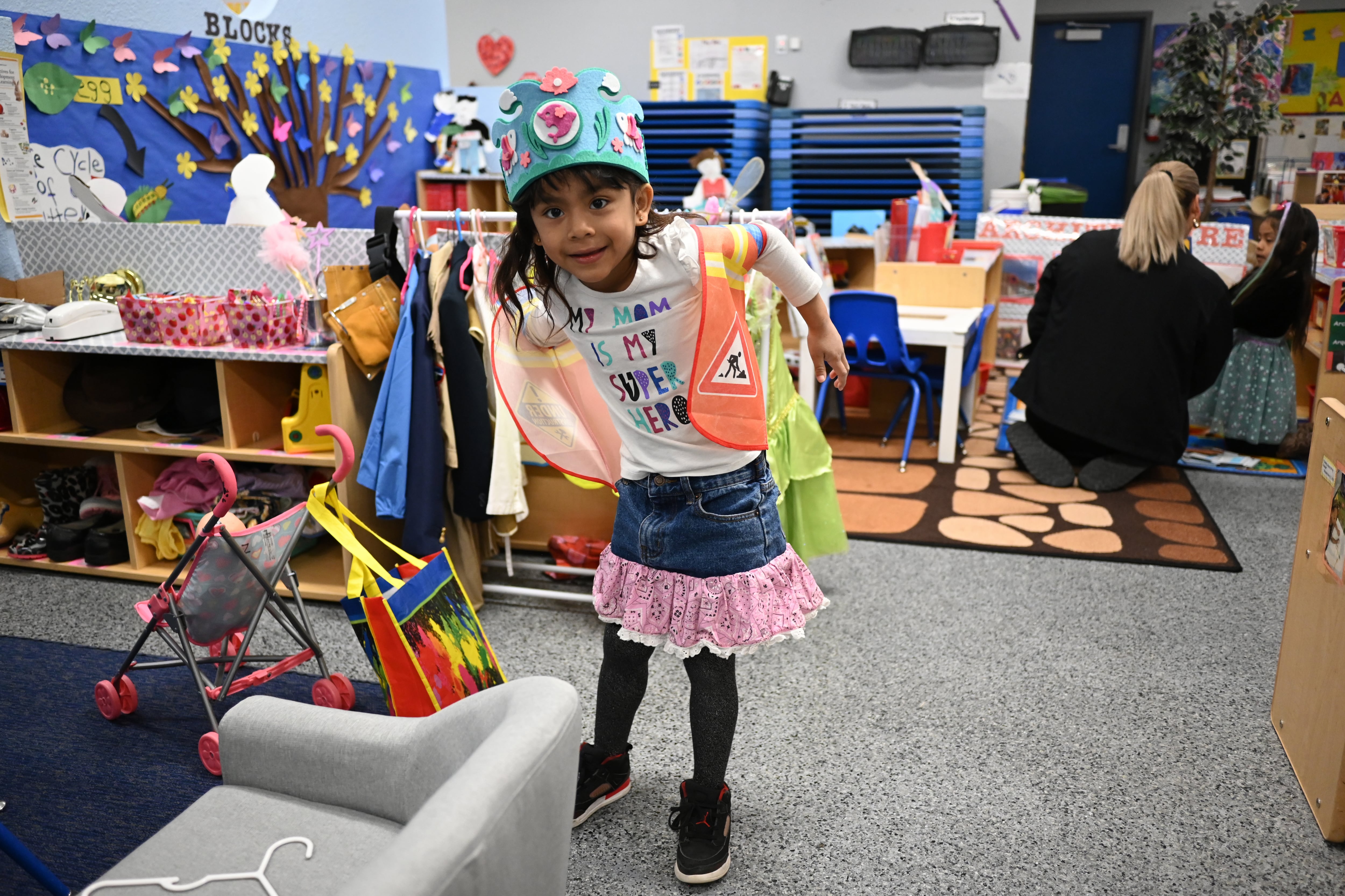 Una niña se viste con un chaleco de seguridad en su clase para jugar. Su playera blanca dice “Mi mamá es mi superhéroe” en ingles.