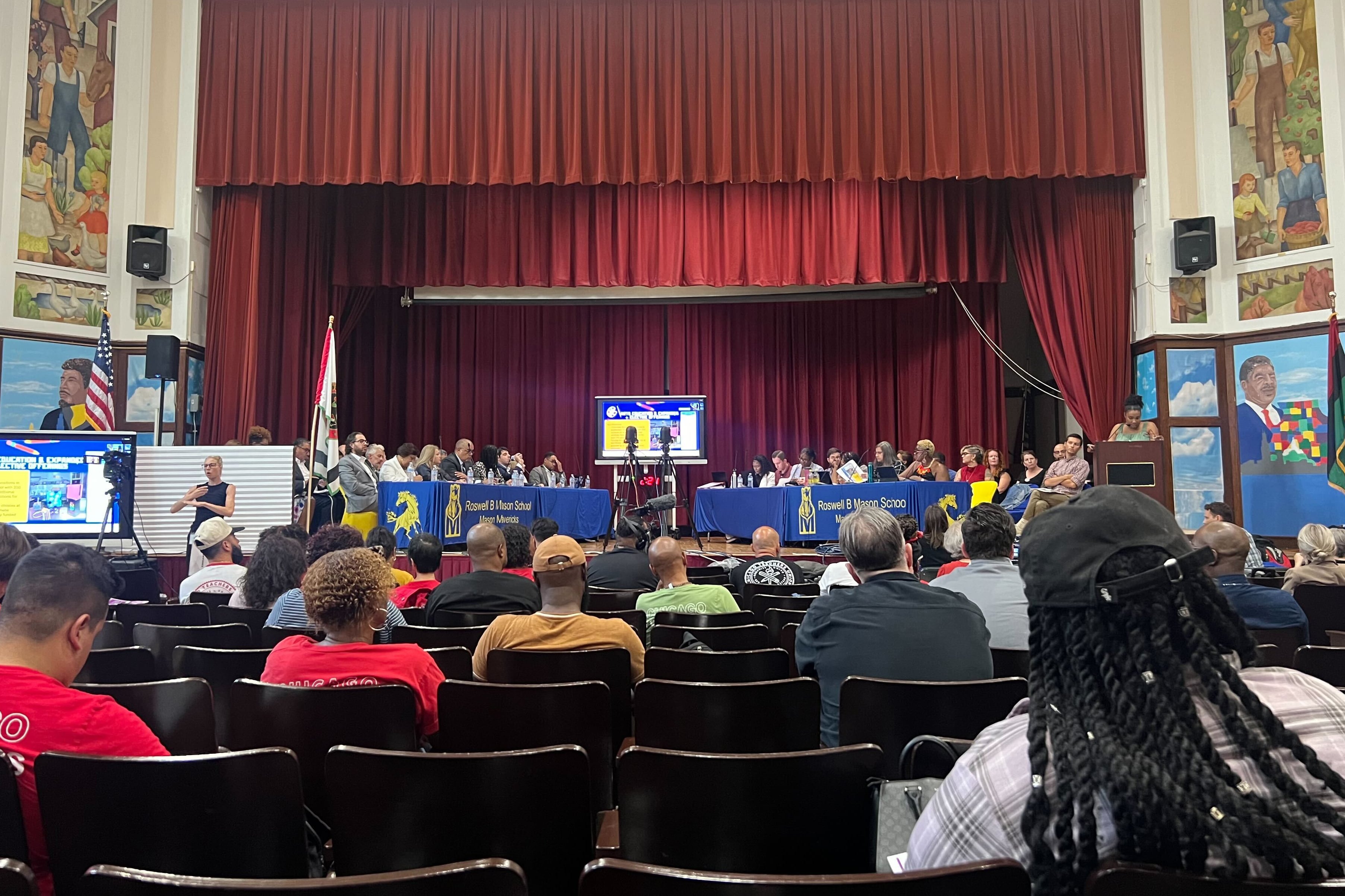 A large group of people sit at tables in an auditorium with large red curtains in the background and people sitting in seats in the foreground.