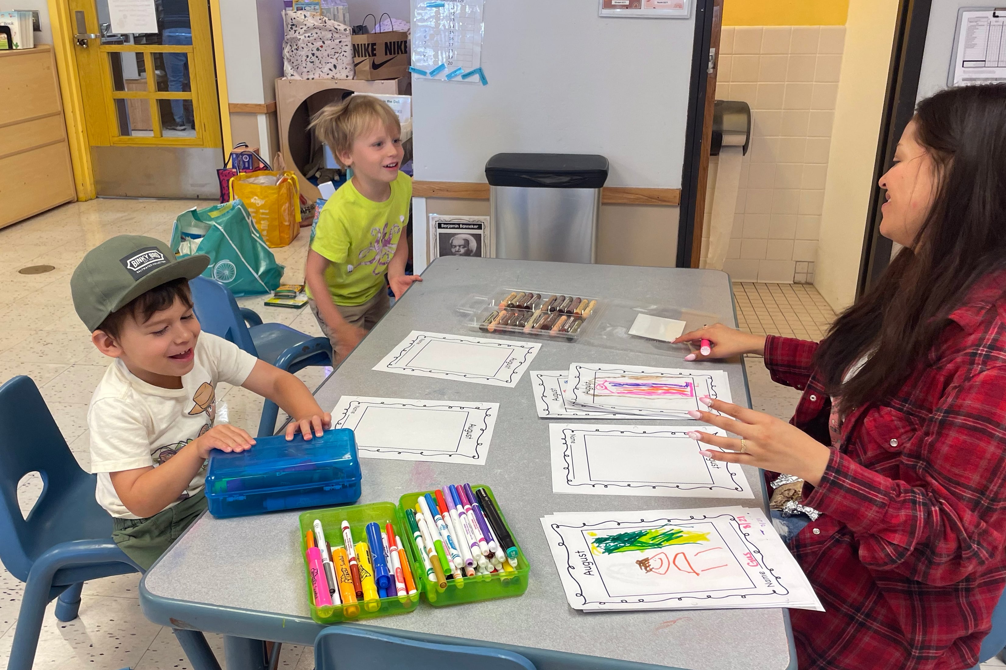 Two young students sit at a table with a teacher while they work on a class assignment.