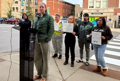 A woman in a green jacket stands at a podium in front of people holding signs.