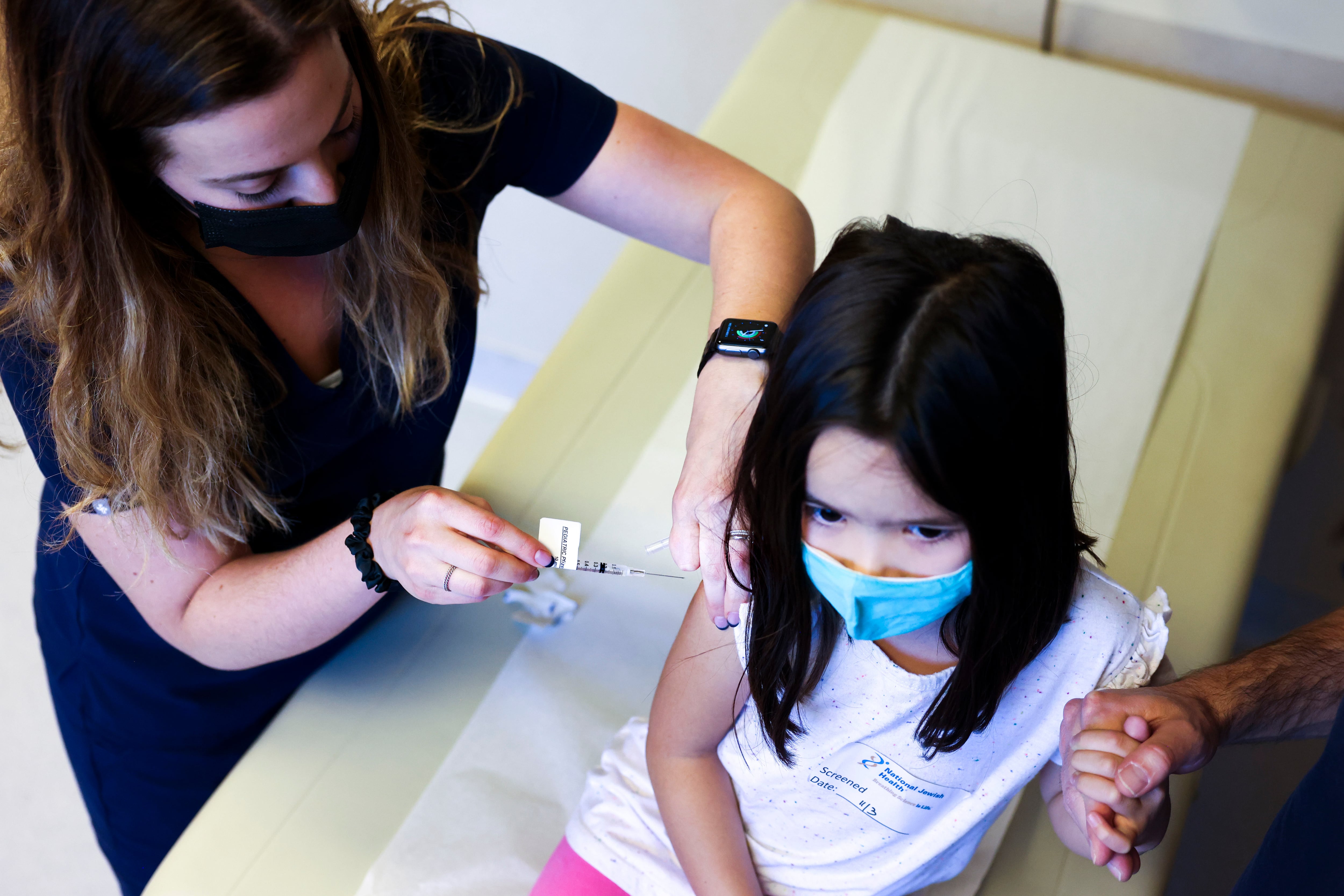 A nurse, wearing dark-colored medical scrubs and a black face mask, vaccinates a young girl while her father holds her left hand.