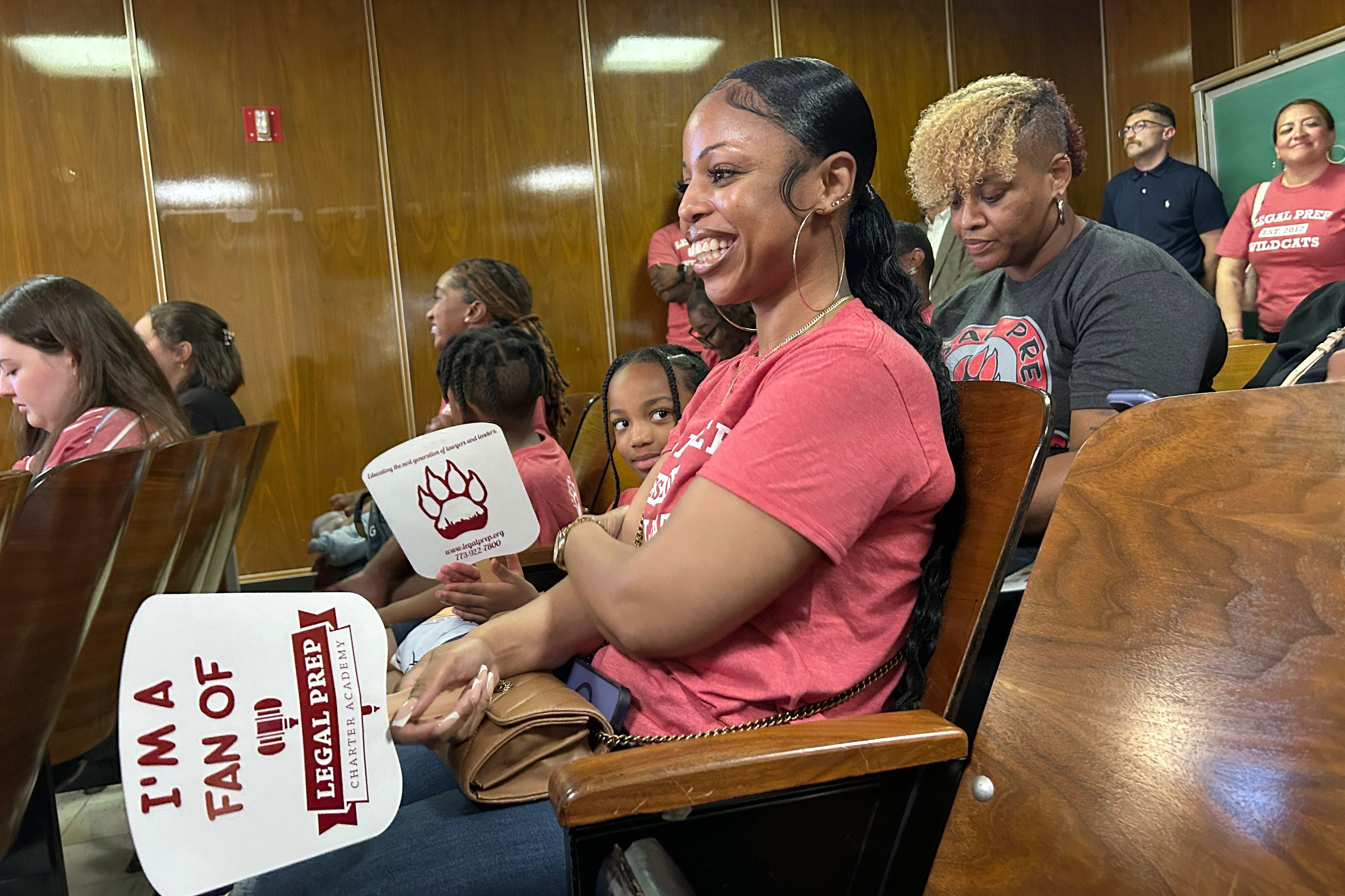 A woman in a pink top holds a fan surrounded by others in the audience at a meeting.
