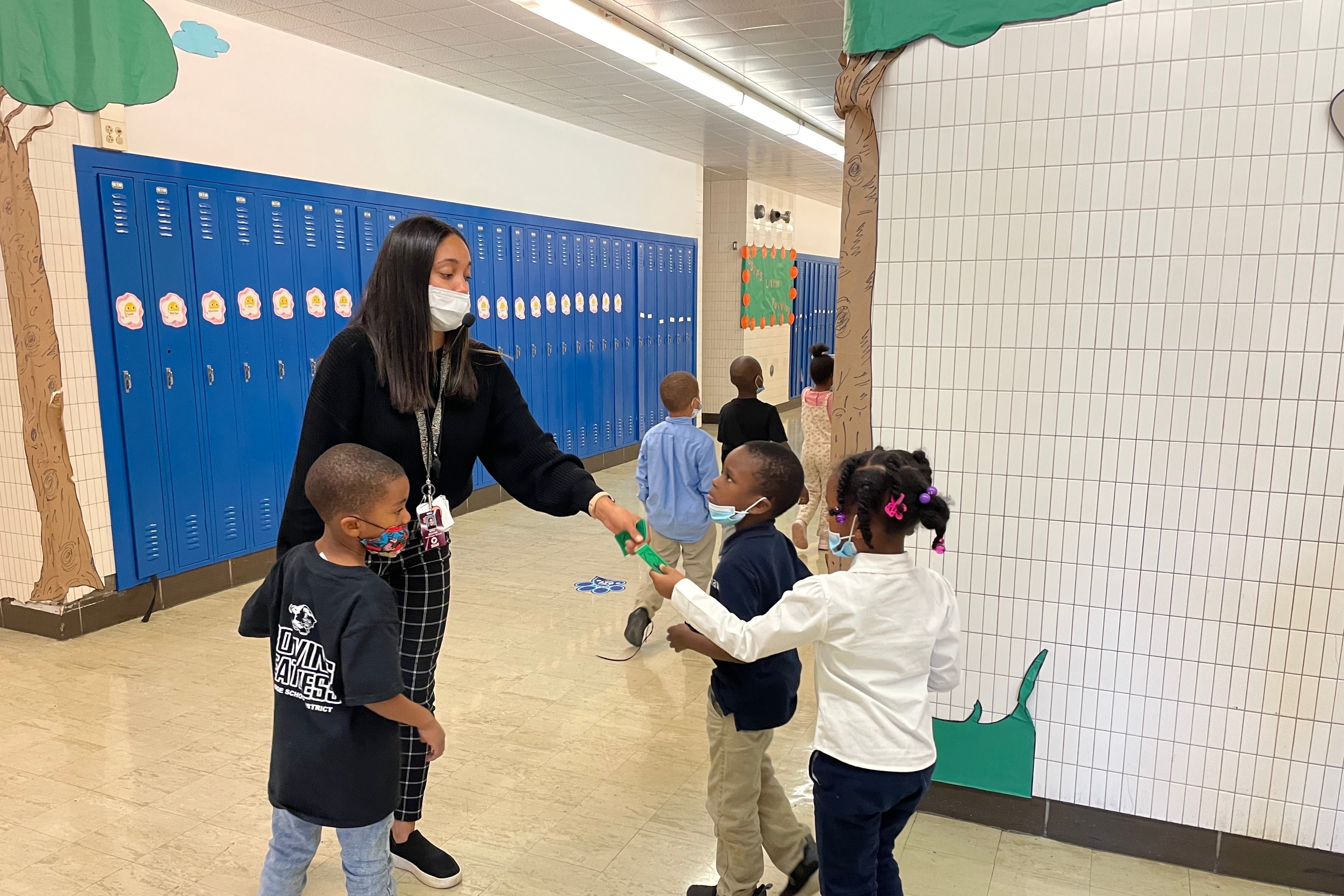 Kindergarteners hand small green slips to their teacher in the hallway, which has blue lockers and drawings of trees on the corners of white tiled walls.