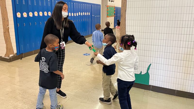 Kindergarteners hand small green slips to their teacher in the hallway, which has blue lockers and drawings of trees on the corners of white tiled walls.