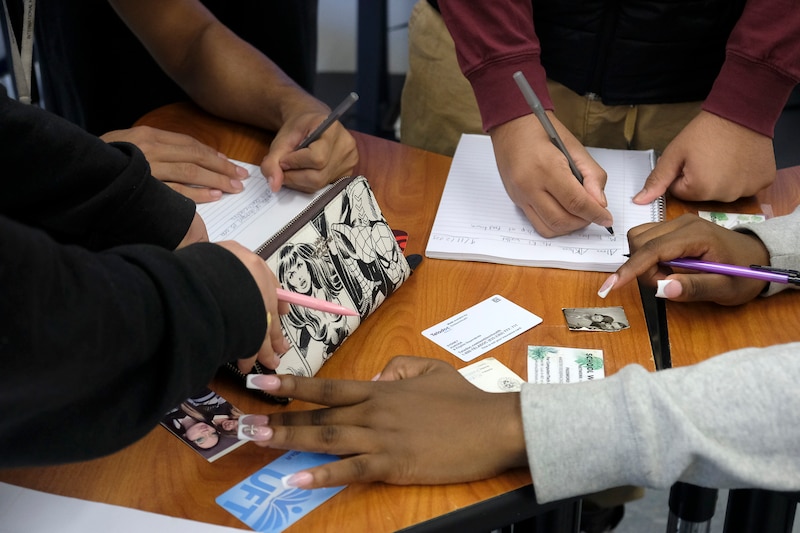 A photograph of four high school students' hands all writing on paper on wooden school desks.