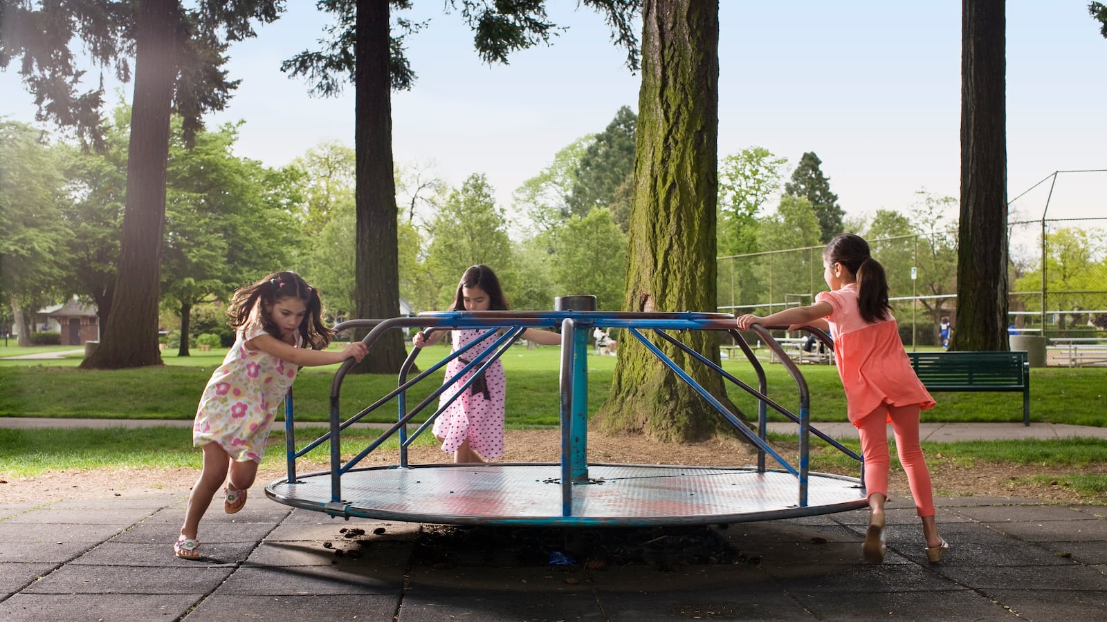 Three girls pushing a metal merry-go-round at the playground.