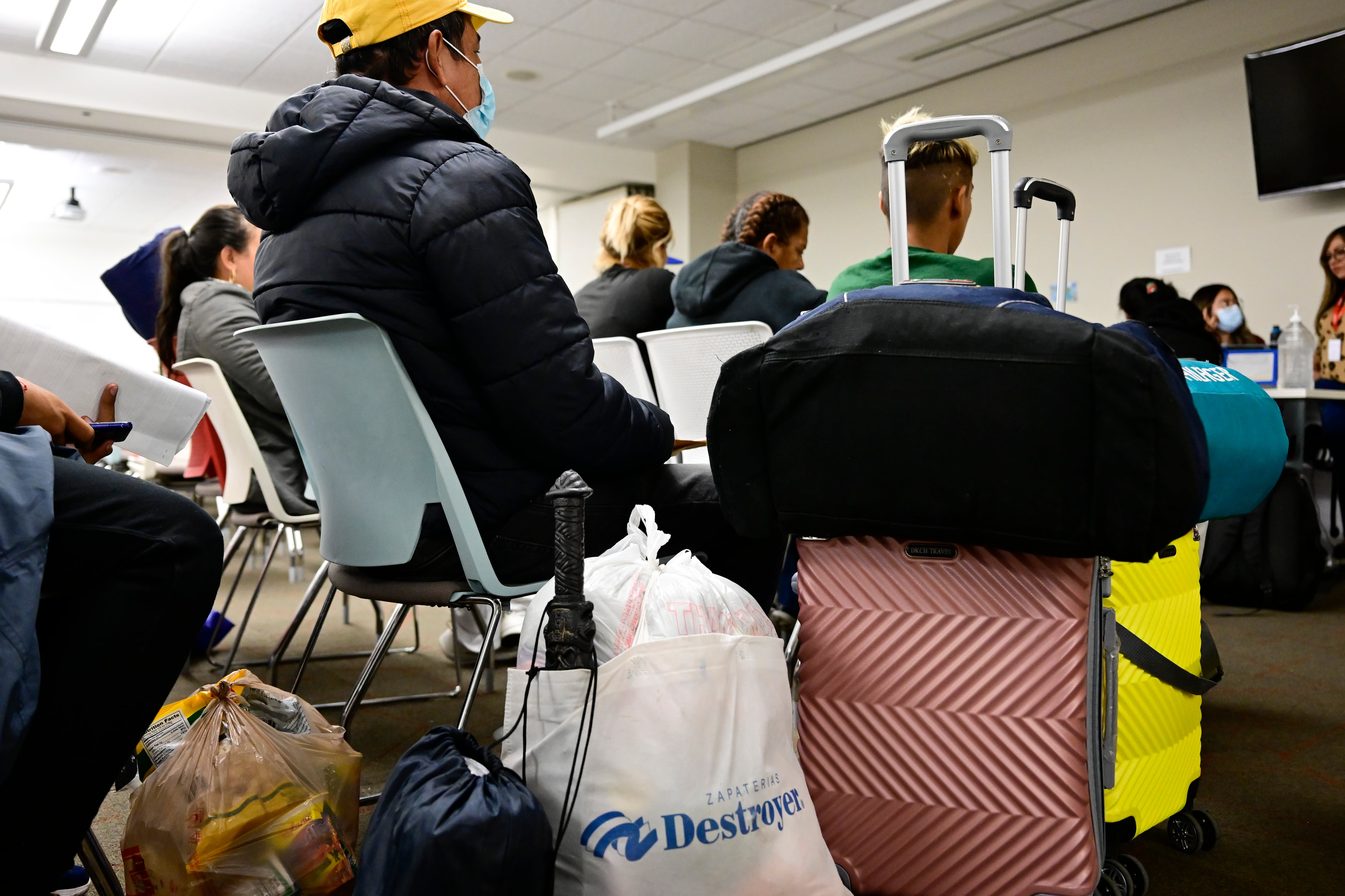 Bags of different sizes and a pink suitcase sit in front of a person sitting in a chair wearing a black jacket and a yellow cap. People sit in chairs waiting in the background.