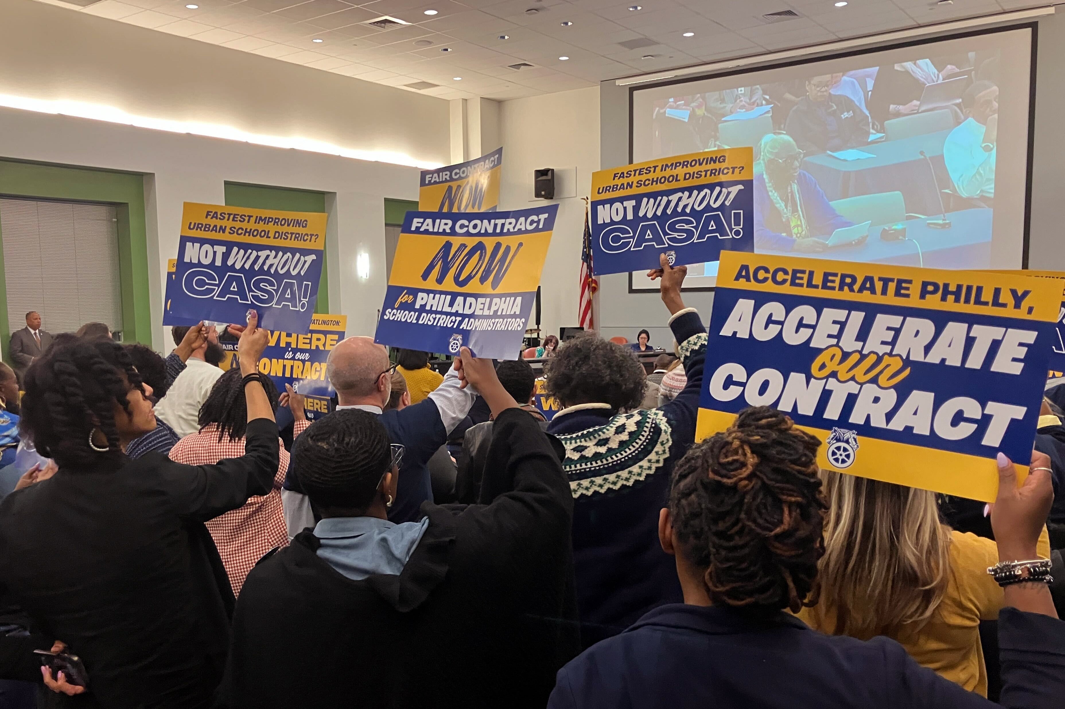 A photograph of a meeting room full of adults, some of them are holding up protest signs.