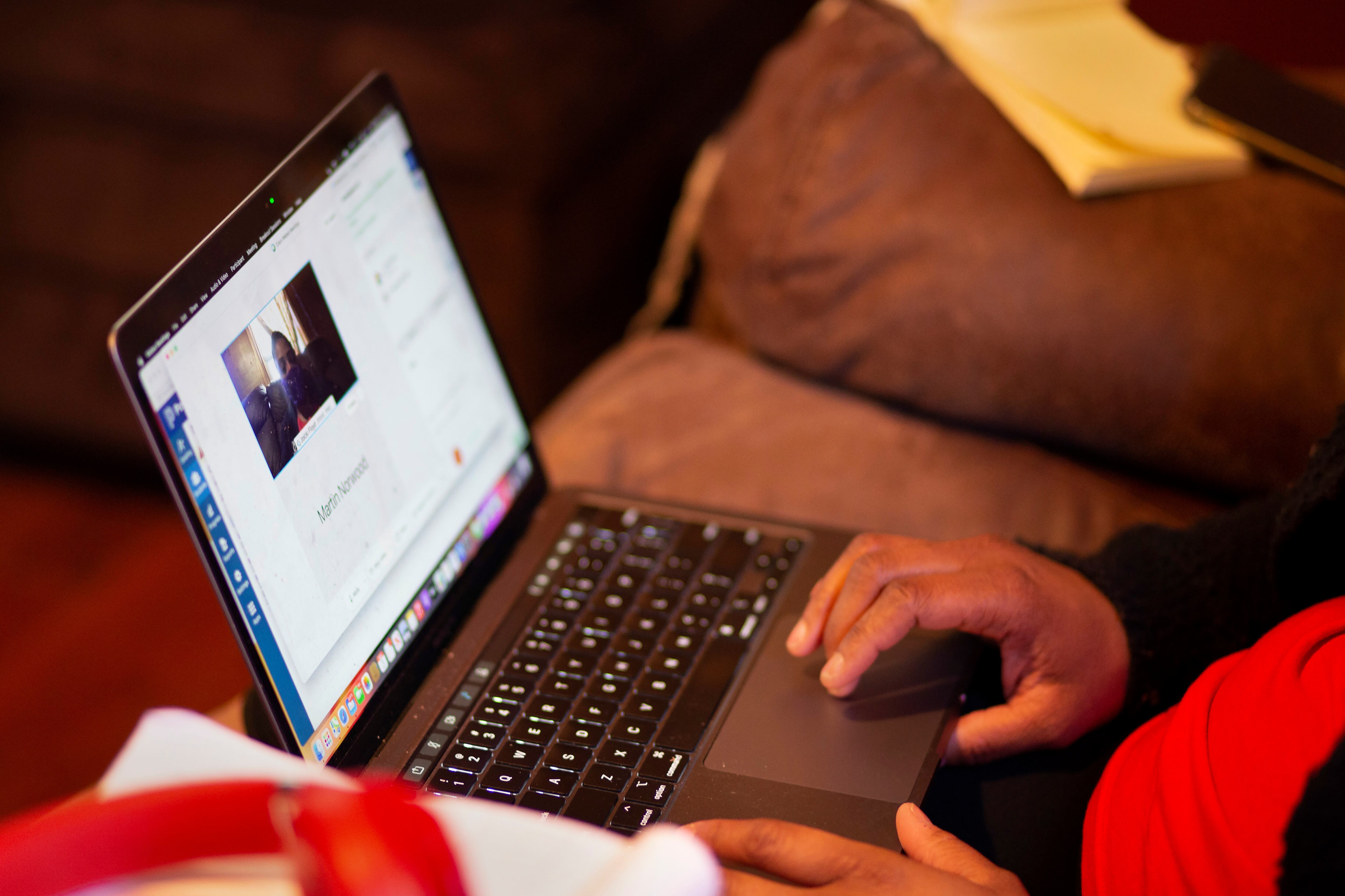 A close up shot of a laptop with someone’s hands on it. The person, not fully pictured, is sitting on a leather couch.