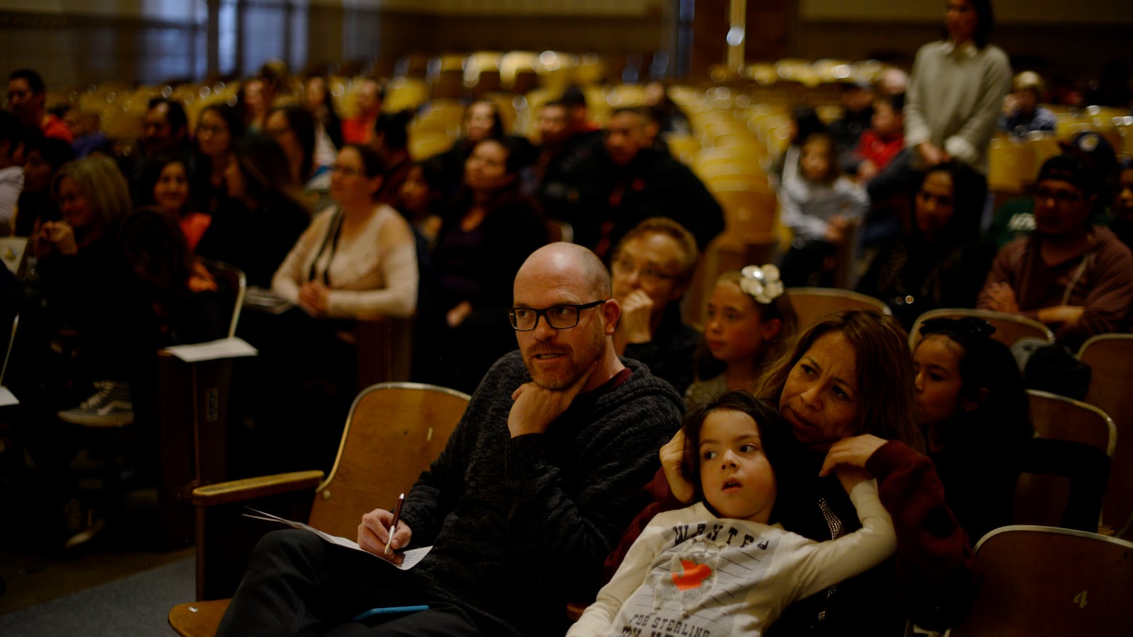 Parents and some children sit in a school auditorium as students compete in a spelling bee.