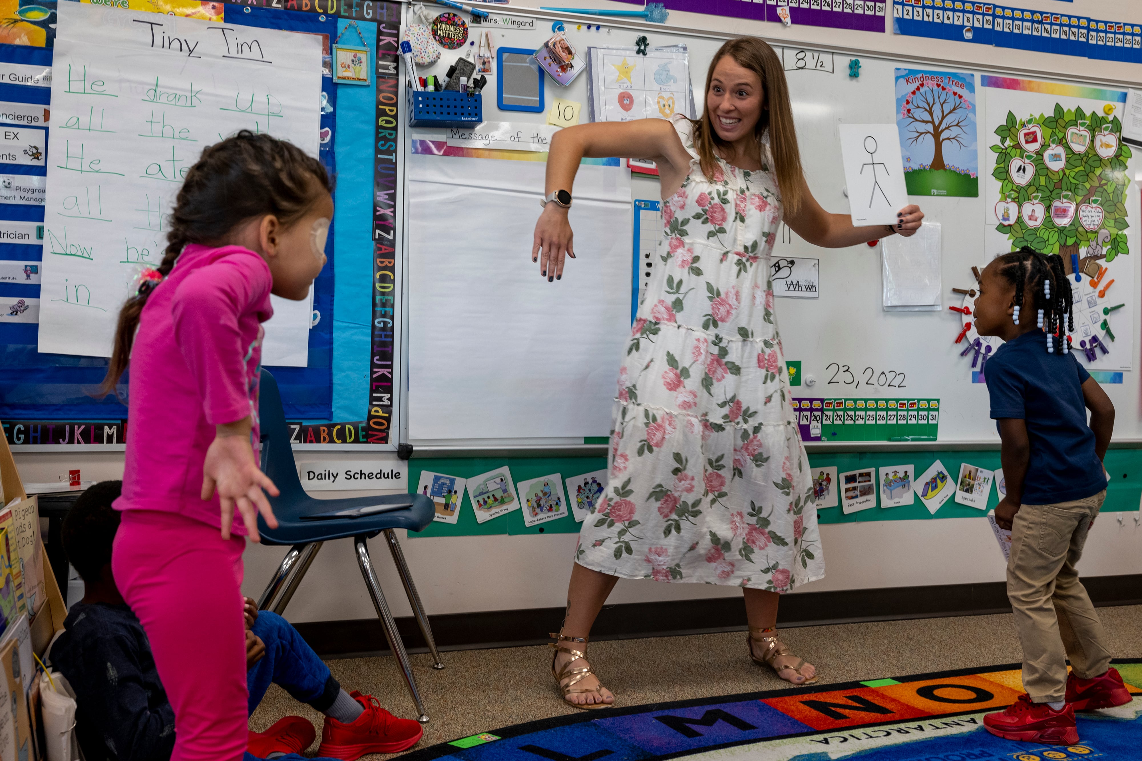 A young woman in a flowered dress dances with two young girls in a preschool classroom.